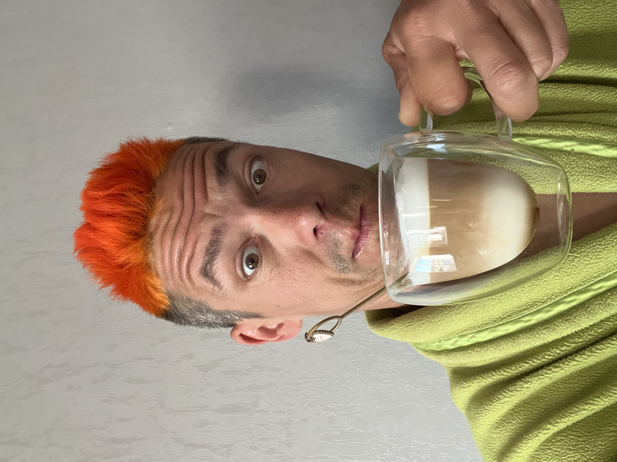 A man with orange hair is holding a clear glass mug filled with a layered beverage, possibly coffee with milk. He's wearing a green fleece jacket. The man has light skin, brown eyes, and is looking directly at the camera with a surprised expression. A small, ornate spoon is visible near his ear. The background is a plain, light gray wall. The location, Chaikovsky, Russia, cannot be verified from the image.