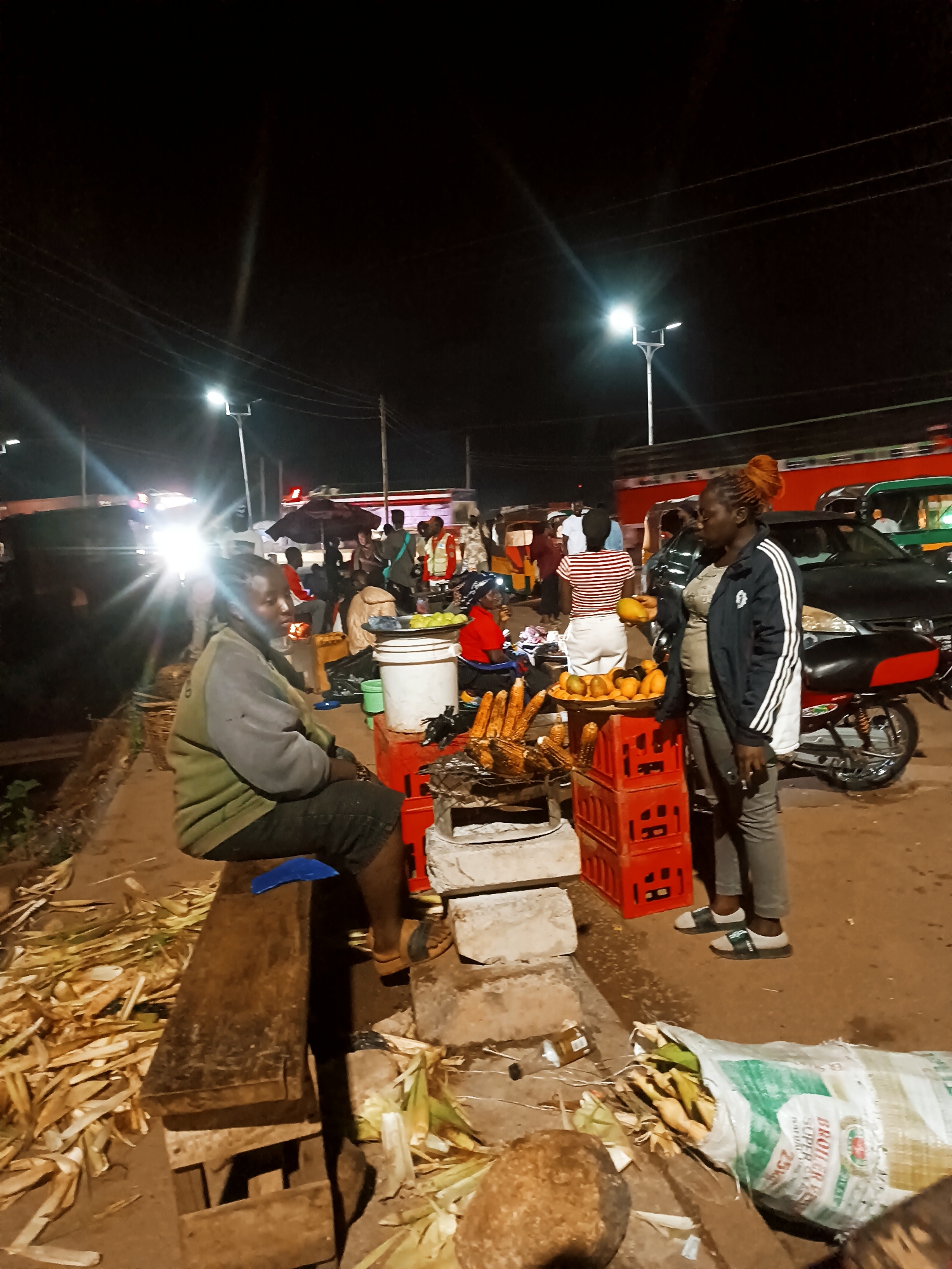 This image depicts a bustling night market scene, likely along a street or informal trading area. The setting is dark, illuminated by bright streetlights and vehicle headlights, which cast strong lens flares across the frame.
In the foreground on the left, a woman wearing a light green/grey sweatshirt and dark shorts sits on a rustic wooden bench, looking towards the right. Numerous corn husks and cobs are scattered on the ground around the bench, indicating active processing or consumption of corn.
Central to the scene, a vendor's stall features a metal grill over hot coals, cooking several corn cobs. Above the grill, a tray of green fruits is displayed, and below it, stacked red plastic crates serve as a stand. Another tray containing yellow/orange fruits (possibly mangoes) is placed beside the grilled corn.
To the right of the stall, a woman with reddish-orange hair, wearing a black jacket with white stripes and grey pants, stands facing left. She holds a yellow fruit in her hand. Behind her, a motorcycle is parked, and a dark car is visible, along with other people in the background, suggesting ongoing market activity. A person in a red and white striped shirt is also seen further back.
Several vehicles, including a car with bright headlights and what appears to be a bus or truck, are present in the busy background. Power lines stretch across the top of the dark sky. On the ground in the lower right, a large white sack filled with corn and husks is visible, bearing partial text "BROILER FEED" and "25KG." The ground appears unpaved or dusty, strewn with husks and some debris like a plastic bottle. The overall atmosphere conveys a lively and informal nocturnal trading environment.
