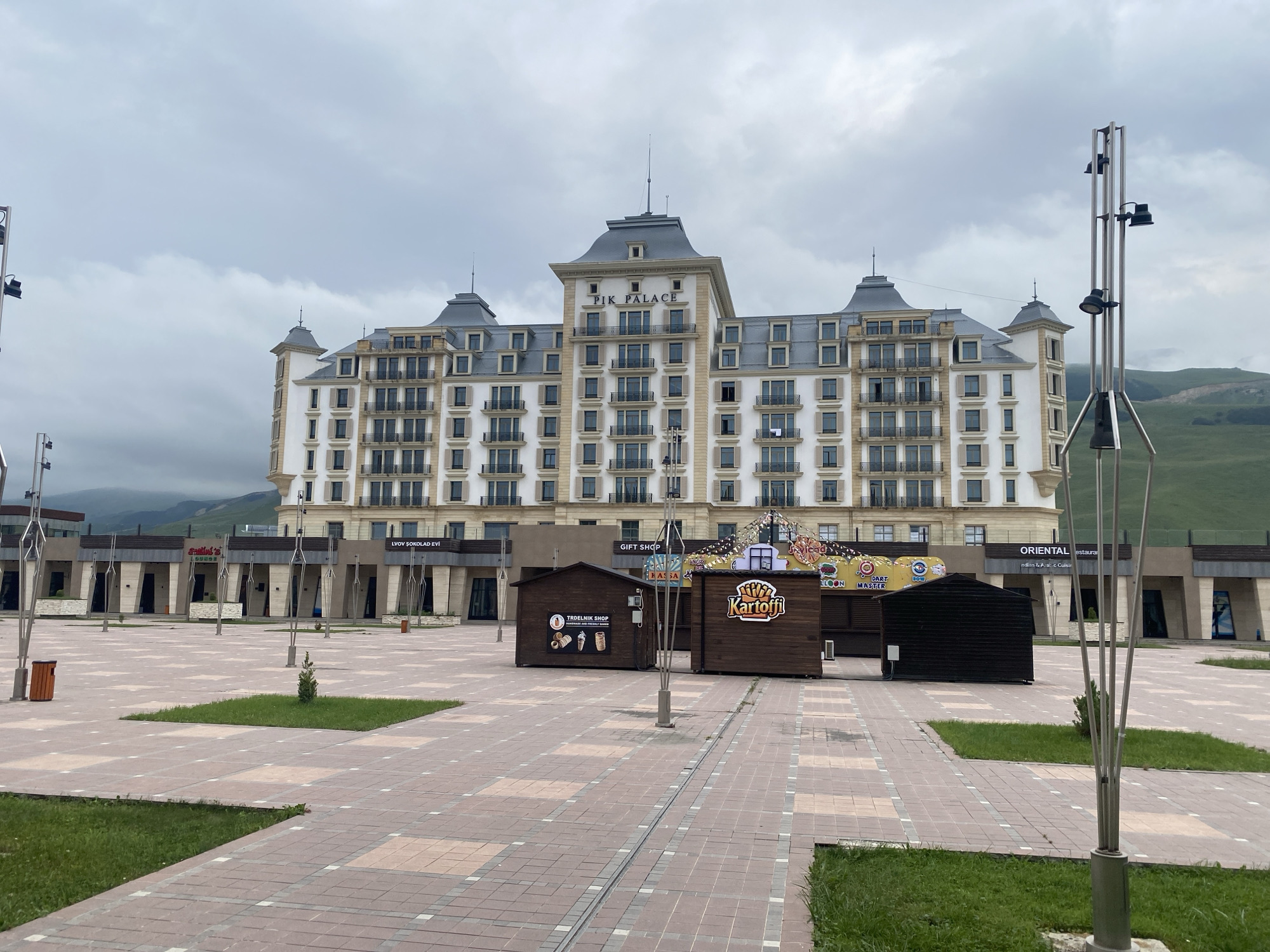 The image depicts the "Pik Palace" hotel in Kuzun, Azerbaijan, under a cloudy sky. The grand hotel, with its white facade, multiple balconies, and distinctive grey roof, dominates the background. In front of the hotel, a plaza is paved with reddish-brown and grey square tiles, interspersed with small patches of green grass and occasional young trees. Several low-lying commercial buildings line the plaza, featuring various shops and kiosks. One kiosk is labeled "Kartoffi," and another appears to be a "Gift Shop." Tall, modern streetlights with multiple lamps stand at intervals across the plaza. In the distance, rolling green hills are visible, suggesting a mountainous or hilly terrain surrounding the town. The scene is devoid of any people.