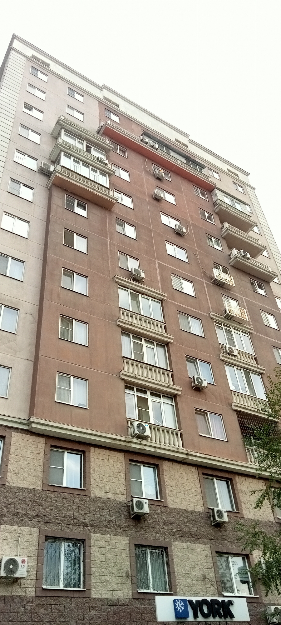 A wide-angle, low-angle, vertical shot captures the facade of a multi-story apartment building in Almaty, Kazakhstan. The building is primarily clad in a reddish-brown plaster, with a lighter beige stone veneer on the lower floors. Numerous windows of various sizes are visible, many with white frames, and some featuring decorative balconies with balustrades. Several air conditioning units are mounted on the exterior walls. A sign with a snowflake logo and the word "YORK" is visible near the bottom of the frame. The sky is overcast and appears bright white. The image is focused on the architecture and design of the building.