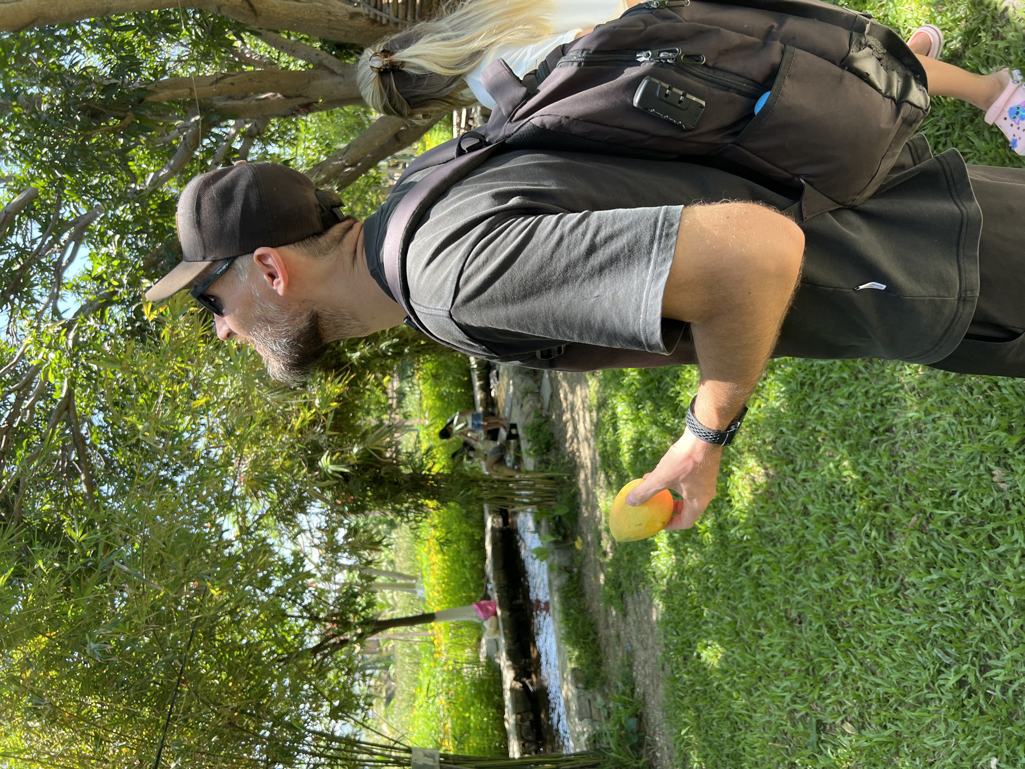 An outdoor scene in Nam Cam Ranh, Vietnam, depicts a man holding a fruit and looking upwards into a tree canopy. The man, positioned in the mid-ground right, wears a dark t-shirt, dark shorts, a reversed dark baseball cap with black sunglasses pushed onto it, and a dark backpack. He has a beard and a watch or bracelet on his left wrist. In his left hand, he holds a yellow-orange, ovoid fruit. He is looking directly upwards into the dense green foliage of a large tree. To his right, partially obscured, is a second individual with long blonde hair tied back with a claw clip, wearing a white top and pink Crocs-style footwear. In the background, lush green vegetation includes bamboo stalks, a manicured grass lawn, and a narrow stream with stone banks flowing left to right. Beyond the stream, fields of yellow flowers and more greenery are visible. Two small figures are partially visible near the stream in the far background. A pink bag rests on the stream bank. The scene is brightly illuminated by natural sunlight, indicating a clear day.