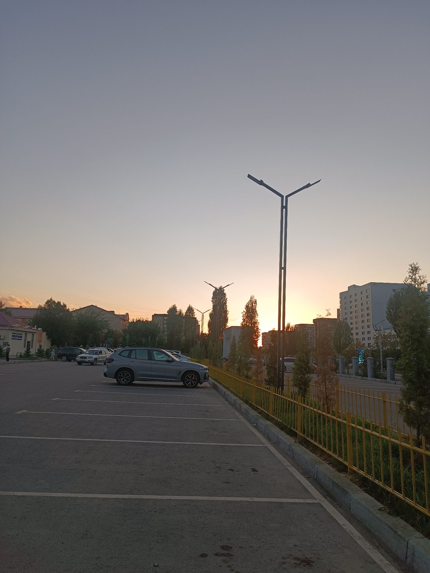 A wide-angle, low-angle shot captures a parking lot with a grey SUV parked in the foreground, facing left. The parking lot is paved with asphalt and marked with white lines. A yellow metal fence runs along the right side of the parking lot, with trees and buildings visible behind it. In the background, the sky displays a soft gradient of colors, indicating either sunrise or sunset. Tall streetlights with multiple arms stand out against the sky. Further back, several residential buildings and trees are visible. The scene appears to be in an urban environment, likely in Bishkek, Kyrgyzstan, given the typical architecture and the presence of streetlights. There are no people clearly visible in the immediate foreground, though there are a few figures near the buildings on the left.