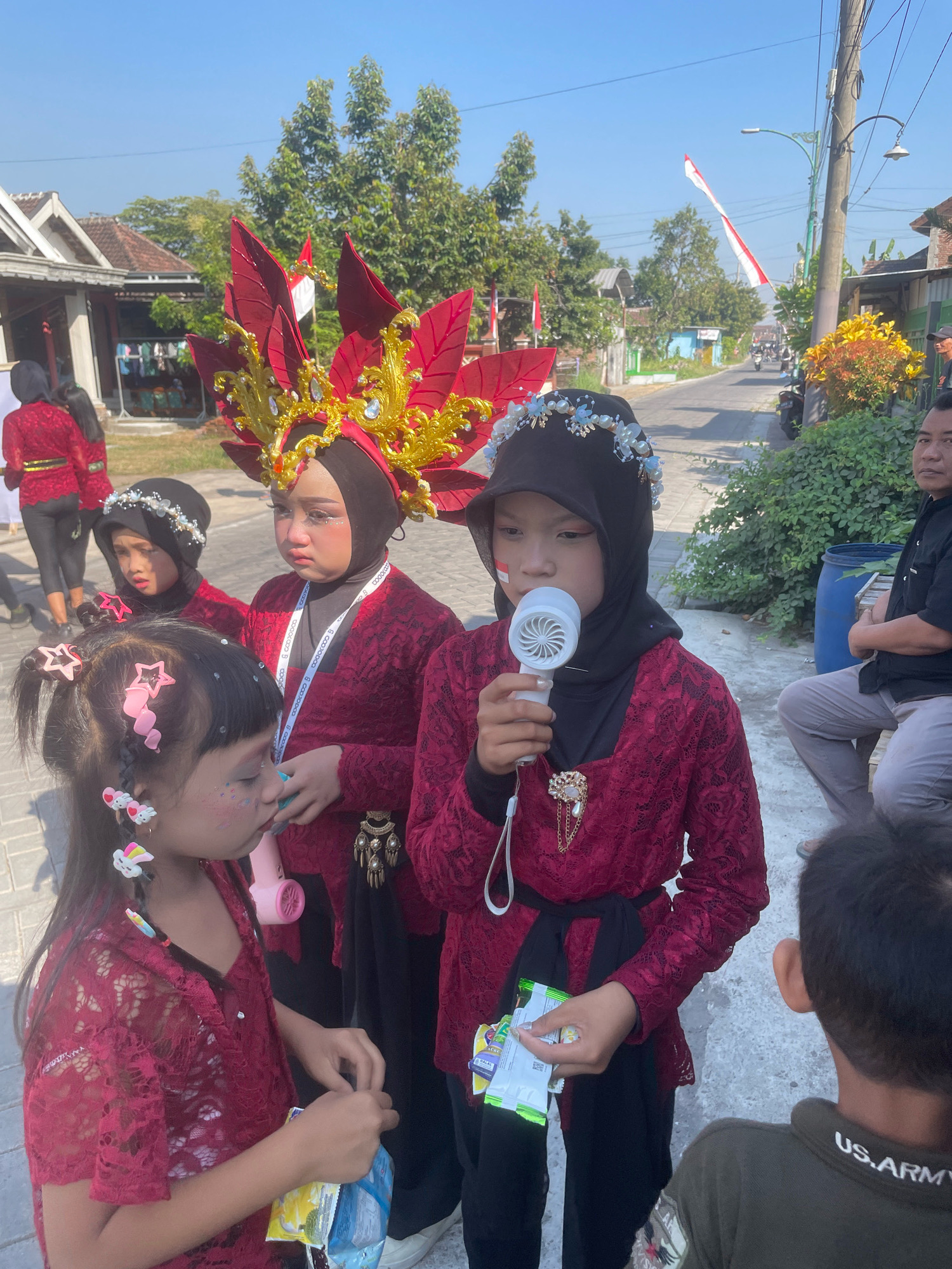In the city of Kebon Waris, Indonesia, on a sunny day, several young girls are dressed in traditional maroon lace clothing with black headscarves. One girl, positioned centrally, wears a large, ornate red and gold headdress adorned with jewels and large red leaf-like elements. She holds a white toy hairdryer to her mouth, which has face paint applied, including a red stripe on her cheek. To her left, another girl also wears a maroon outfit and a black headscarf, with a delicate pearl and floral headband. Her face also shows signs of makeup. A third girl, in the foreground, faces left and is dressed in a similar maroon lace top. She has her hair styled with pink star-shaped clips and bunny-shaped earrings. She is holding a pink toy hairdryer and a packet of snacks. Behind them, other individuals are visible in traditional attire, and in the background, a street scene unfolds with buildings, trees, and a few motorcycles. The Indonesian flag is visible flying on a pole. A man in a black shirt is seated to the right, observing the scene. The overall environment suggests a cultural event or performance taking place outdoors in a residential area.