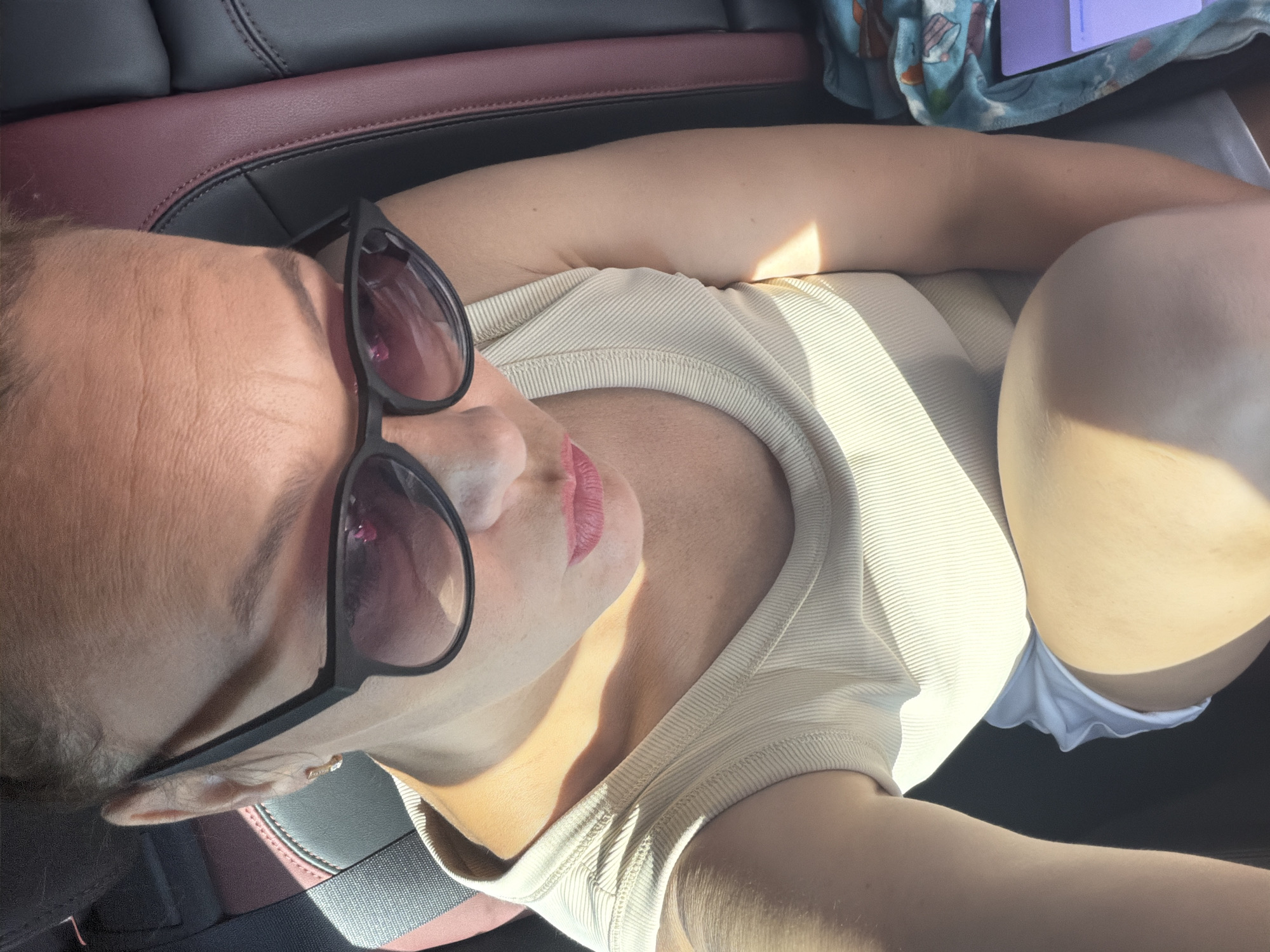 The image shows a close-up, selfie-style shot of a woman wearing a beige tank top and white shorts. She is seated in a car, with the headrest of the driver's seat visible in the background. The woman is wearing black sunglasses and has pink lipstick on. Sunlight is casting shadows across her face and body. A patterned blanket and a tablet device are visible on the right side of the frame. The woman is looking down and to her left. There are no discernible landmarks or city features visible that would indicate a specific location within Chaikovsky, Russia.