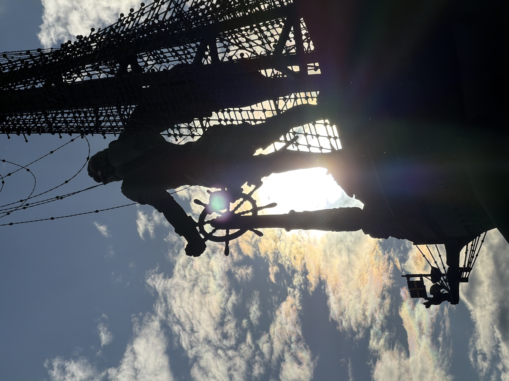 The image shows a low-angle, upward view of a ship replica. The ship's rigging and a statue of a person holding the ship's wheel are silhouetted against the sky. The sky is blue with textured white clouds, with the sun appearing to be behind the statue. The location is the replica of the Santa Maria in the city of Columbus, Ohio.