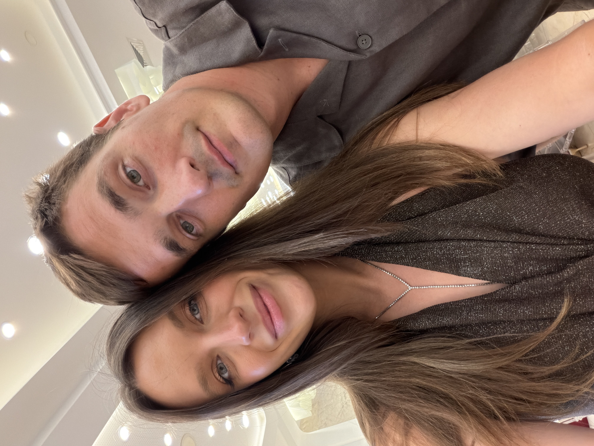 A close-up selfie shows a man and a woman posing together. The woman is positioned in the foreground on the left, with her head tilted towards the man. She has long, brown hair and is wearing a shimmering dark dress with a V-neckline, adorned with a delicate diamond necklace. Her eyes are a striking blue, and she has a gentle smile. The man is to her right, his head resting on her shoulder. He has short brown hair and is wearing a dark grey collared shirt. His eyes are also a light shade of green or hazel. They appear to be indoors, possibly in a brightly lit room with ceiling lights visible in the background. The overall impression is one of intimacy and closeness. The scene is not identifiable as being in Nikšić, Montenegro.