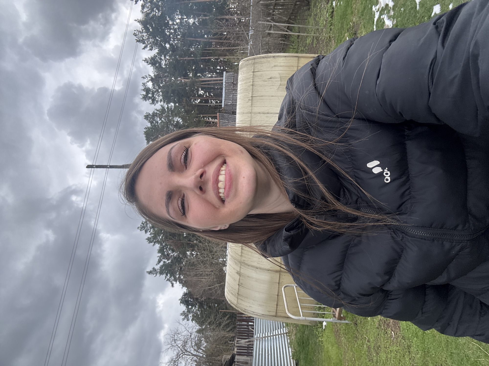A young woman is centered in the frame, smiling and facing the camera. She is wearing a black puffer jacket. The background includes an overcast sky, power lines, trees, and what appears to be a partially visible structure, possibly a shed or outbuilding. The ground shows signs of grass and patches of melting snow. The location is outdoors and could plausibly be in the region of Раздолье, Russia.