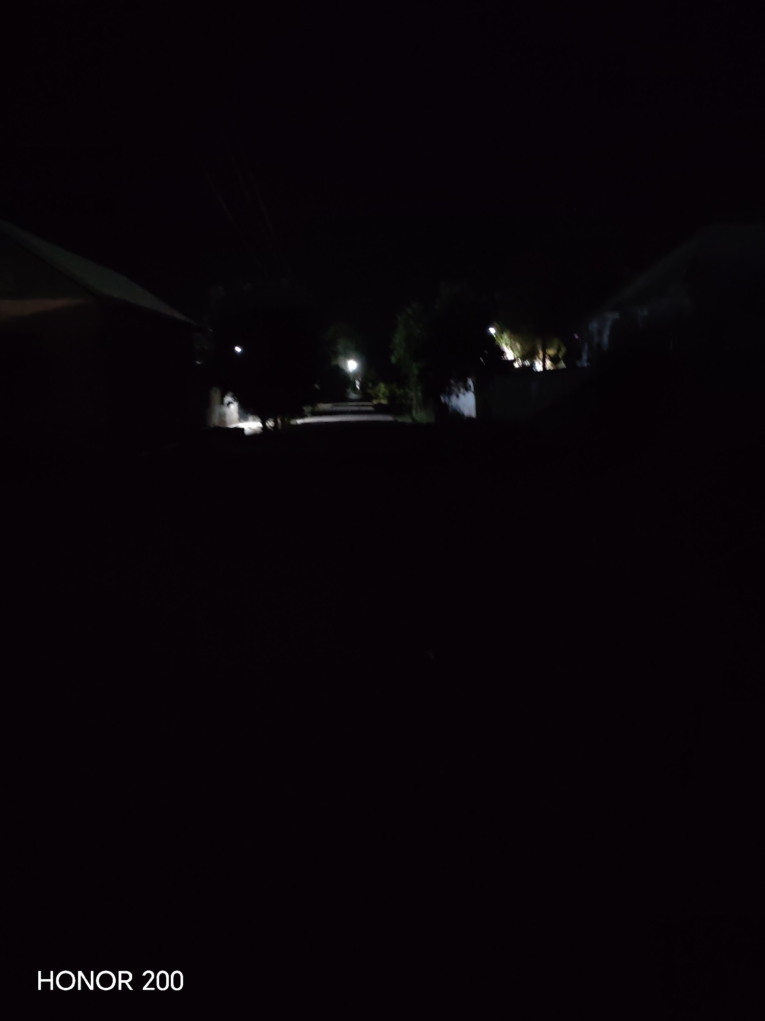 The image is a dark, nighttime scene, likely taken in the city of Kakaydi, Uzbekistan, as indicated by the watermark "HONOR 200" which appears to be a device identifier and not a location. The view is of a street with buildings on either side. A faint light source is visible in the distance, possibly a streetlamp, illuminating a portion of the road. The overall impression is one of low visibility due to the darkness, with the structures and environment being largely obscured. No people or specific actions are discernible.