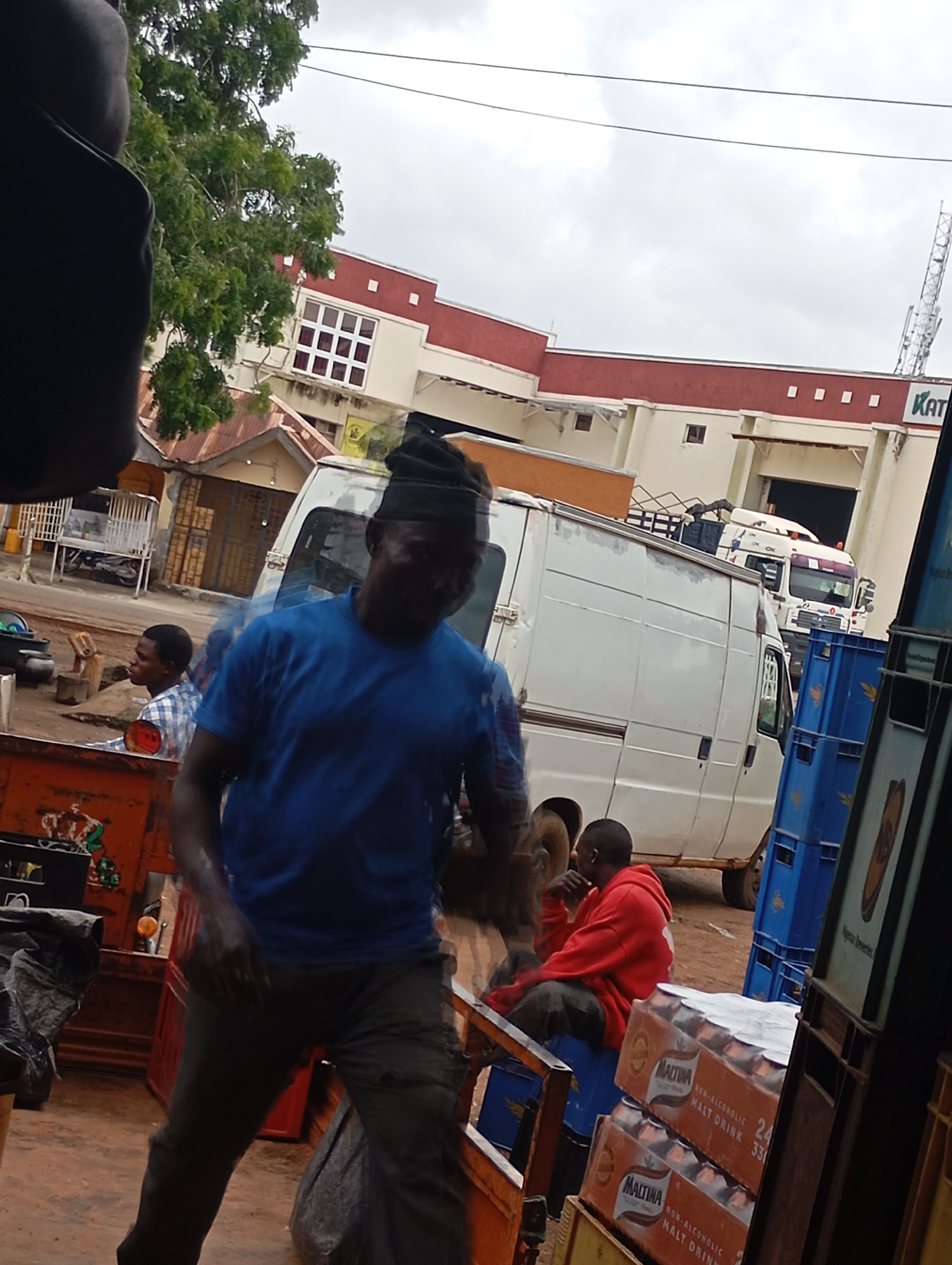 The image depicts an outdoor scene in the city of Shendam, Nigeria, likely near a marketplace or distribution point. In the foreground, a man wearing a blue t-shirt and dark pants, topped with a black cap, is walking towards the right side of the frame. He appears to be in motion, with his body slightly blurred. To his left, another man in a plaid shirt is seated, looking to his left. Further into the background, a white van is parked, and behind it, a white truck is visible. There are stacks of blue plastic crates and orange cases of Maltina non-alcoholic malt drink. In the far background, a large building with a red and white facade is present, along with trees and overhead wires. The sky is overcast.