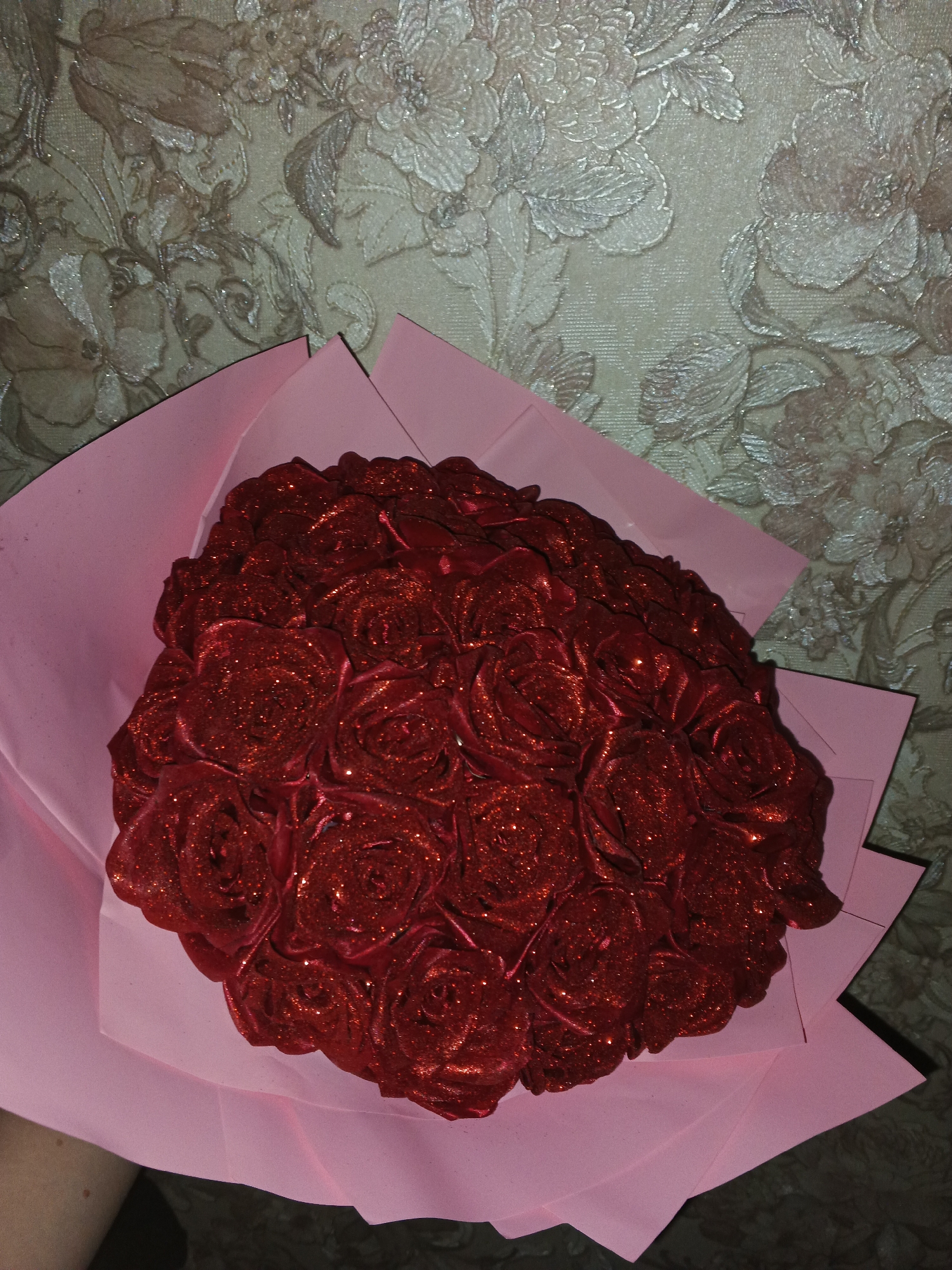 A bouquet of red roses wrapped in pink paper is being held against a wall decorated with floral patterned wallpaper. The roses appear to be made of a textured material and have glitter accents.