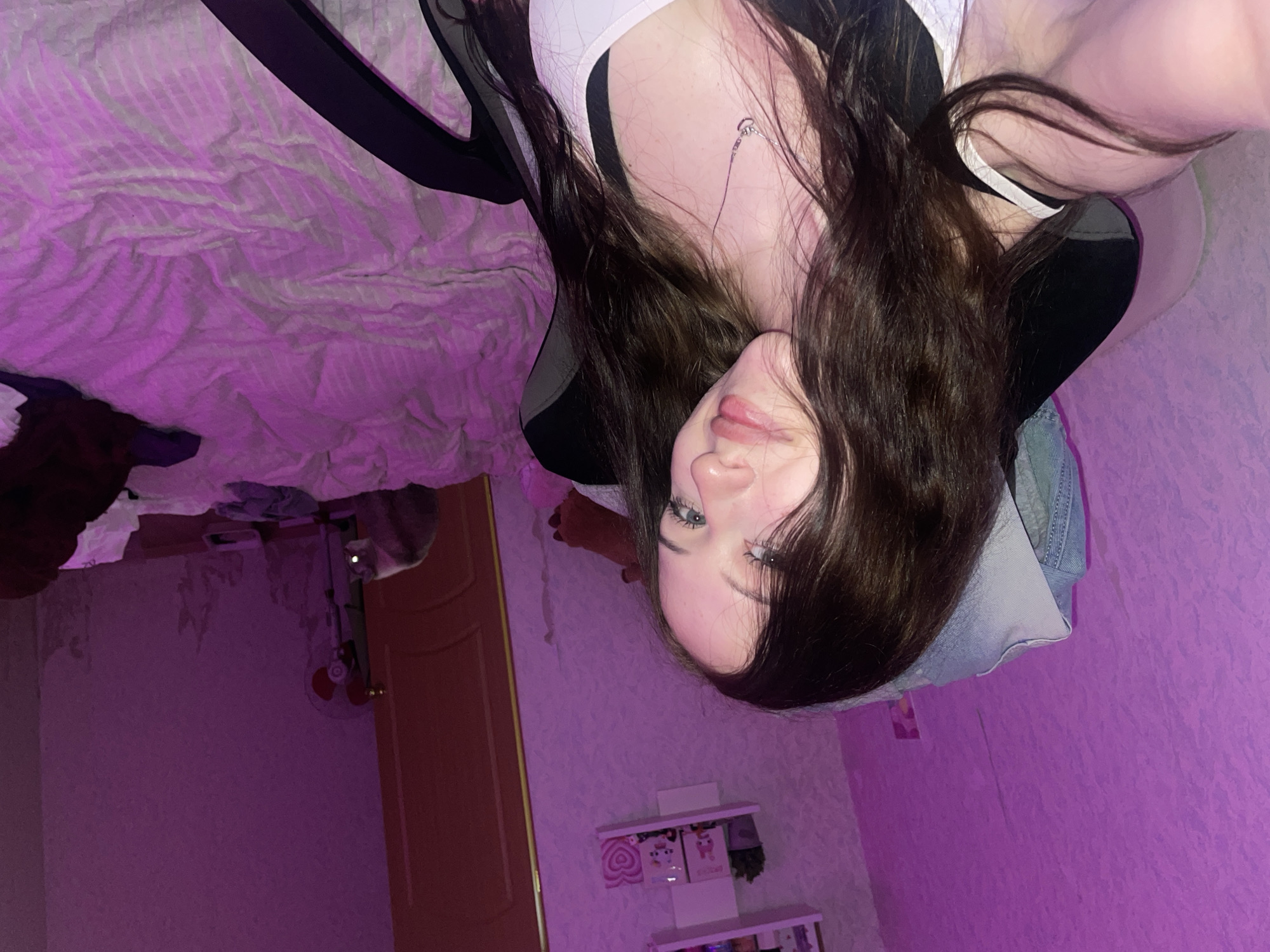 The image is a selfie taken by a young woman with long, dark hair, blue eyes, and a light complexion. She is wearing a white tank top over a black bra and a silver heart-shaped necklace. She is seated in a black gaming chair, with her left arm out of frame, likely holding the camera. The background is dimly lit with pink and purple hues from ambient lighting. To the left, a textured wall is visible, and above it, a white shelf with several small decorative items. To the right, a bed with a pink comforter is partially visible, and a wooden door is slightly ajar. A large, white object, possibly a fan or air purifier, stands near the door. A portion of a duvet cover is draped over a figure in the lower right quadrant of the image. The scene appears to be a bedroom. There is no information in the image to suggest a specific location in Votkinsk, Russia.