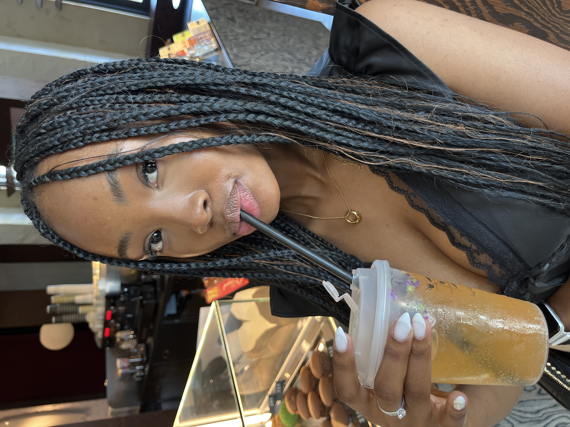 A woman with dark skin and black braided hair is holding a clear plastic cup with a straw and drinking from it. She is wearing a black top with lace trim and a gold necklace. She is also wearing a silver ring on her left hand. The background appears to be a cafe or coffee shop, with a display case containing pastries and a coffee machine visible. The lighting suggests it is daytime. There is no indication of this image being taken in Kazan, Russia.