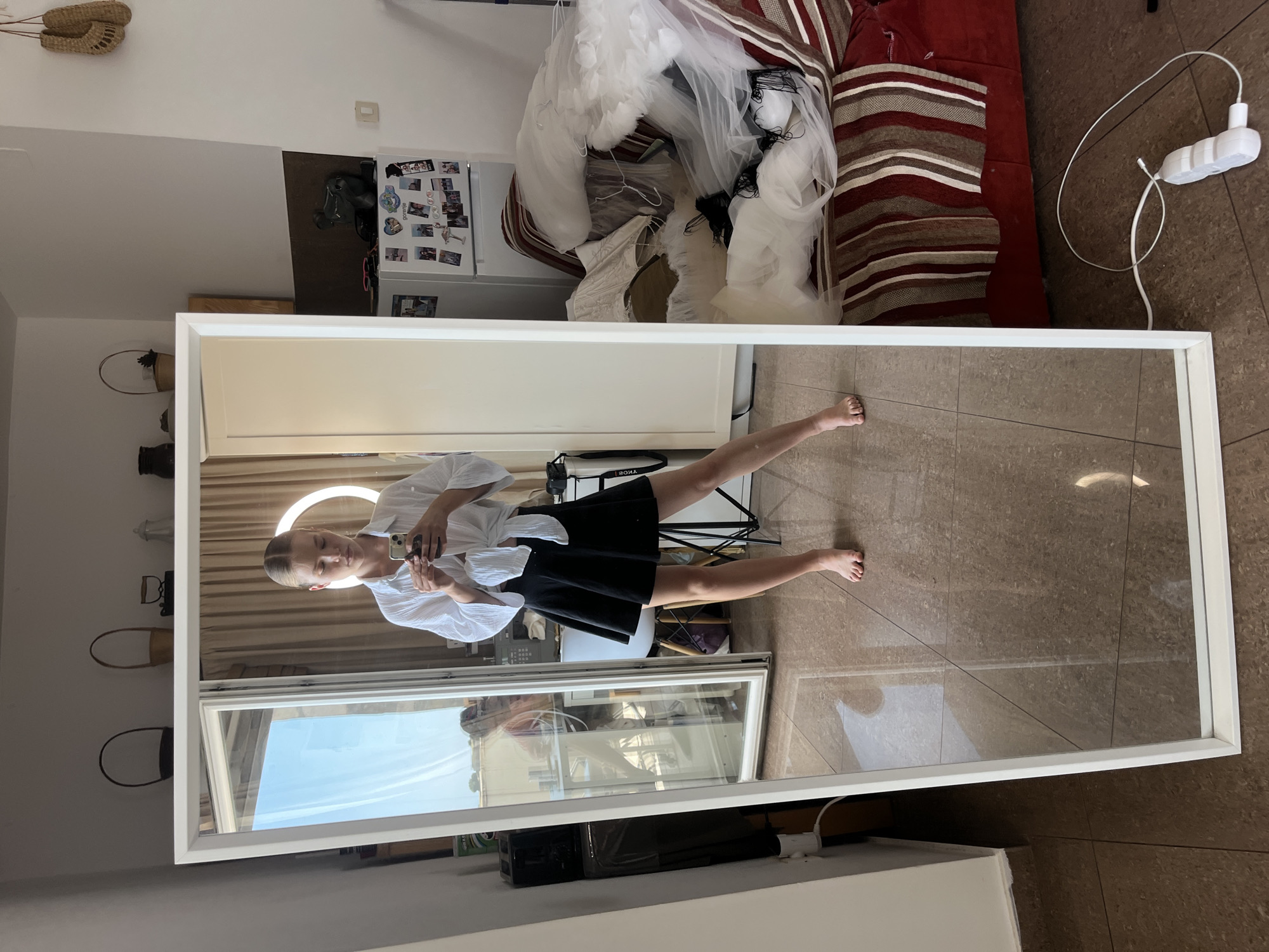 The image shows a young woman taking a mirror selfie in what appears to be a residential room. The woman is wearing a white, cropped, button-down shirt tied at the waist and a black, flared skirt. She is holding a smartphone in her hands and looking at it. Her bare feet are visible, standing on a tiled floor.
Behind the woman, a large full-length mirror reflects the room. The room contains a sofa with a striped cover, piled with white tulle fabric, possibly part of a dress. To the left of the mirror, a refrigerator is visible, covered with various magnets and pictures. There are also baskets on a shelf in the background, and a window showing an outdoor view.
The environment suggests an indoor setting, possibly a bedroom or living area, with a casual and somewhat cluttered atmosphere. The presence of the tulle fabric might indicate an artistic or fashion-related context. There is no explicit indication of the location being in Budva, Montenegro, or any other specific city. The image focuses on the woman and her reflection.