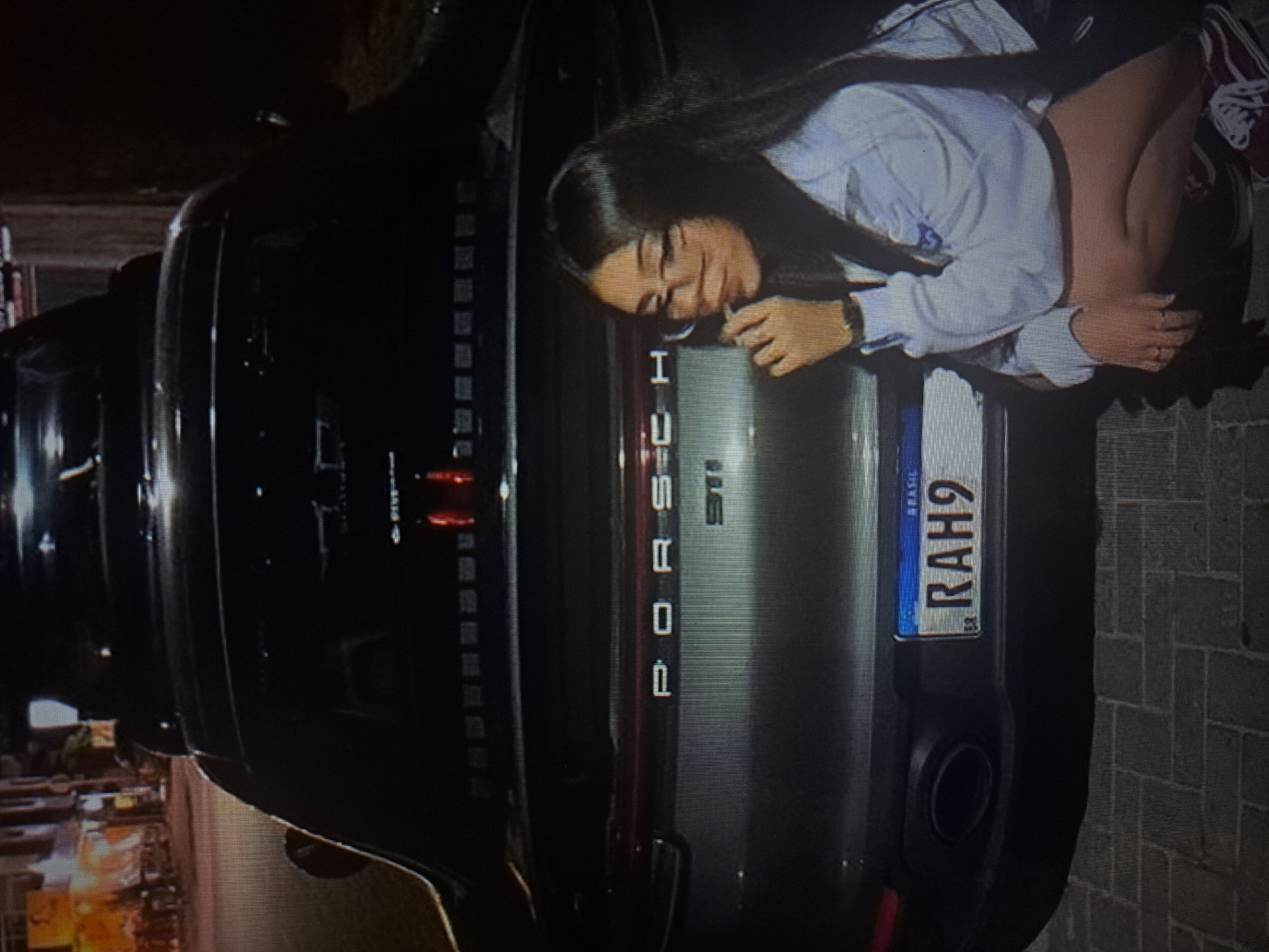 A person is crouching beside the rear of a dark Porsche car. The car’s license plate reads "RAH9" and indicates it is from Brazil. The location is not discernible from the image.