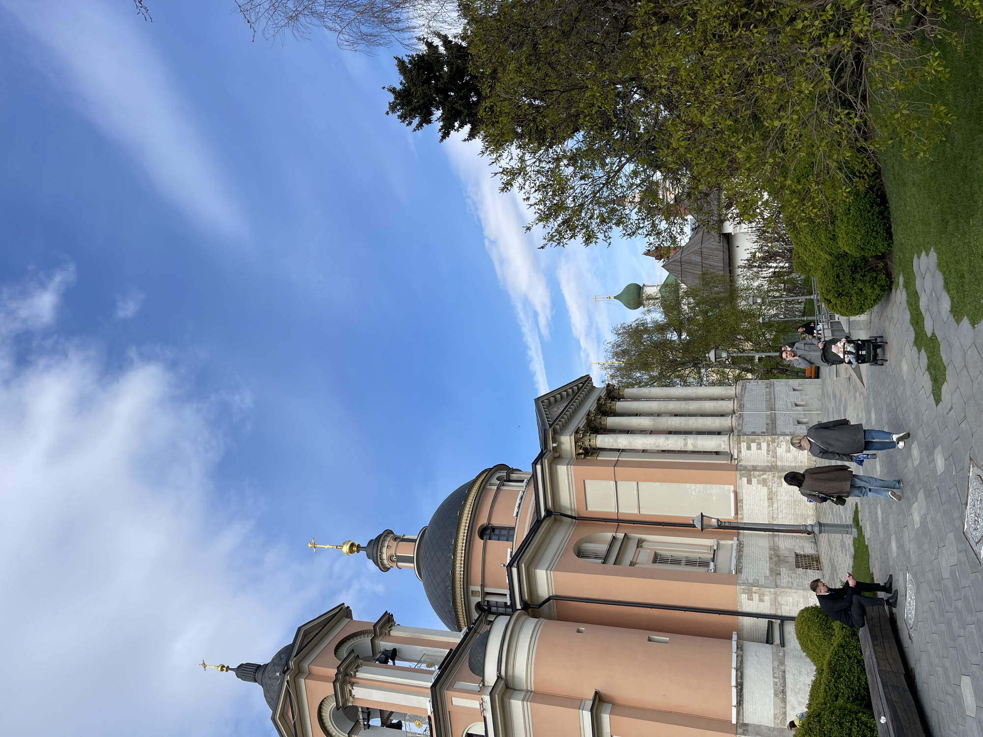 The image captures a view of the Church of St. Catherine the Great Martyr in Zamoskvorechye, Moscow, Russia, under a partly cloudy blue sky. The church has a pale peach exterior, a black dome, and golden crosses atop its structures. Along the right side of the frame, people are present on a paved pathway, including individuals walking and one person sitting on a bench. There are trees and green bushes bordering the pathway. In the background, another building with a green dome is visible.