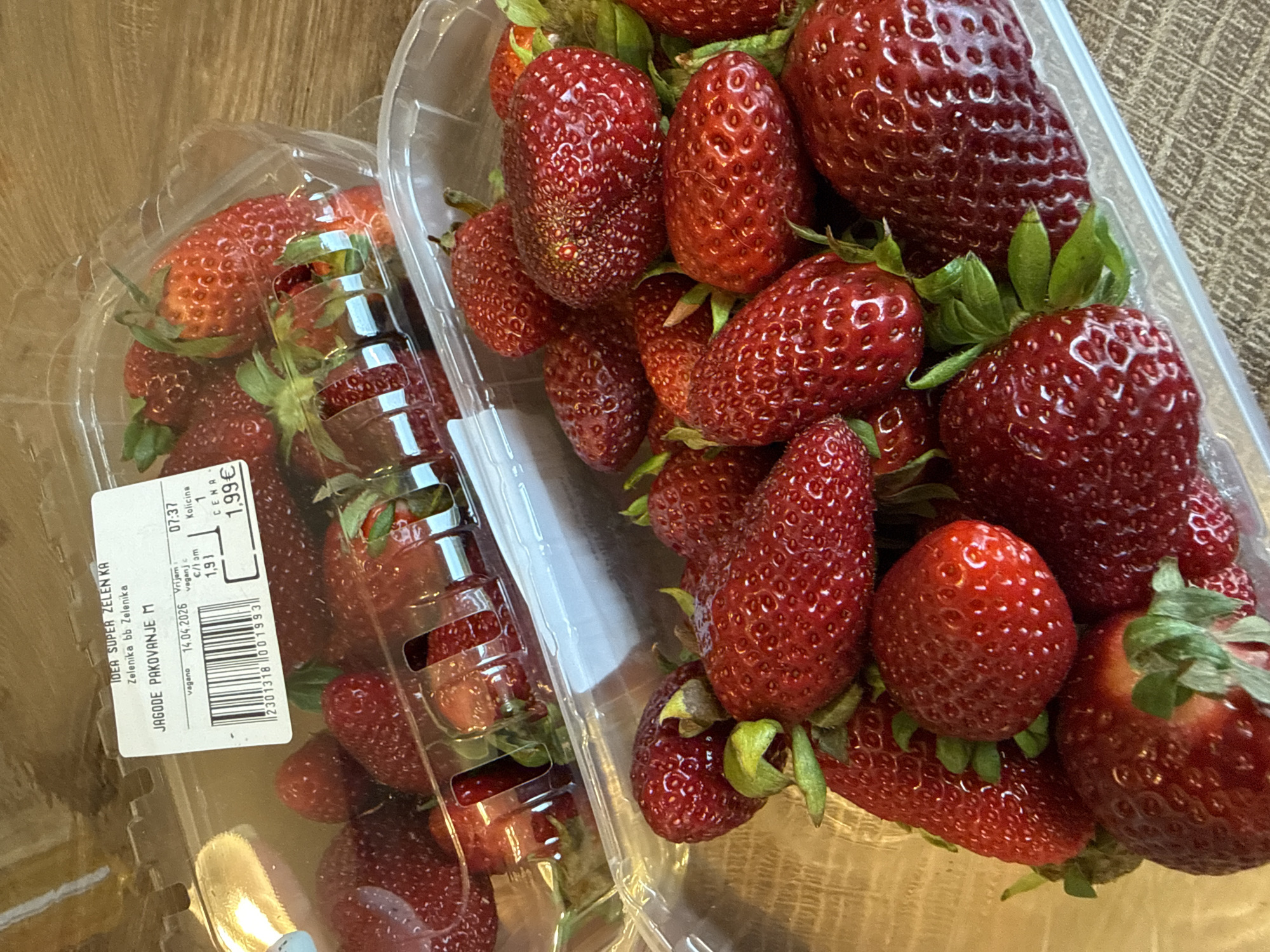 The image shows two transparent plastic containers filled with strawberries. A label on one of the containers indicates the strawberries are "JAGODE PAKOVANJE M" (packaged strawberries M) from "IDEA SUPER ZELEN KA" in Zelenika, Montenegro. The label shows a sell-by date of April 14, 2026, a time of 07:37, a price of 1.99€, and a weight of 1.93 (presumably kilograms). The containers are placed on a wooden surface.