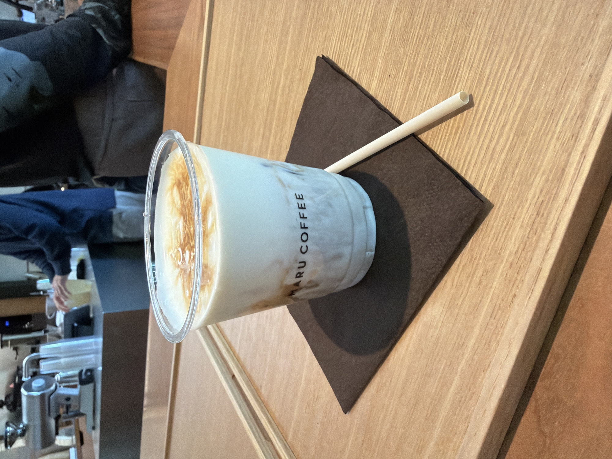 On a wooden surface with natural grain patterns, a plastic cup of a milky beverage with the name "MARU COFFEE" printed vertically on its side rests on a dark brown square coaster. The beverage has a foamy or layered appearance, with darker tones at the top, suggesting a coffee-based drink with milk or cream. A light-colored straw lies next to the cup, partly on the coaster and partly on the wooden surface. In the background, partially visible, is a person seated, wearing a blue jacket and dark pants. There is also an espresso machine with several metallic levers. It is challenging to determine the exact location within New York City without more details.