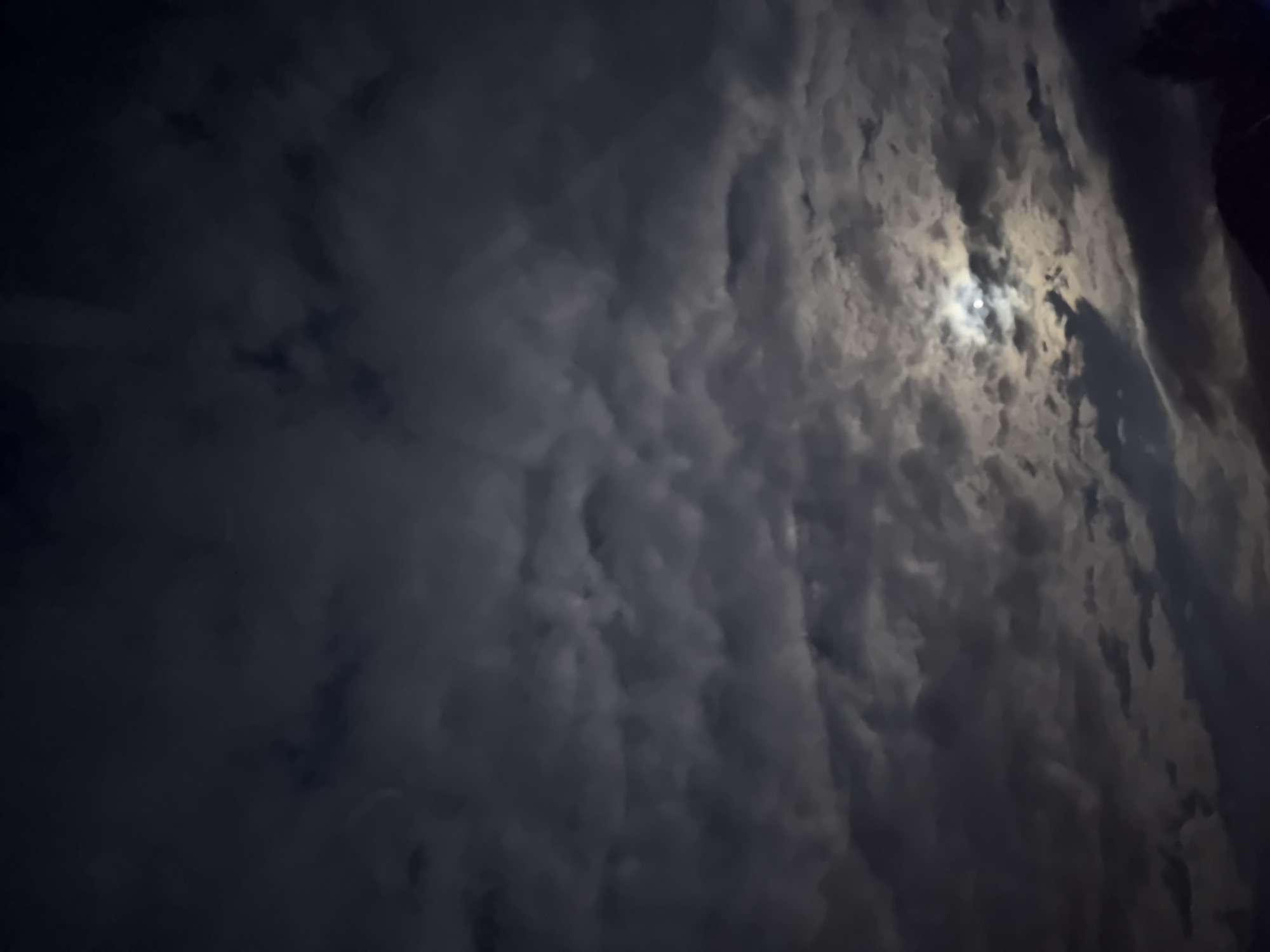 The image depicts a night sky with dense cloud cover in Vrachesh, Bulgaria. A bright, circular object, likely the moon, is visible through a gap in the clouds. The clouds are textured and layered, with varying shades of gray and white. The left side of the image is darker, suggesting deeper cloud cover.