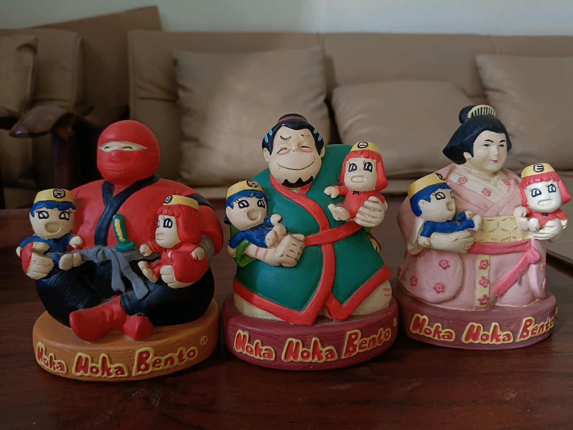 In an indoor setting, possibly a living room, three decorative figurines stand on a wooden surface. The figurines are made of a brightly colored material, likely ceramic or resin. From left to right:
The first figurine is a red ninja-like figure with a red mask covering its face, leaving only eyes visible. It is holding two smaller child-like figures, one on its lap and one held in its right arm. Both children are wearing blue and yellow outfits and have similar facial features. The ninja figure has a green object tucked into its waist, resembling a dagger or sword hilt. This figurine stands on a round base with yellow lettering that reads "Moka Moka Bento".
The second figurine depicts a larger character with a round, smiling face, dark hair, and a goatee. This character is wearing a green robe with a red trim and is holding two child-like figures. One child, on the left, is wearing a blue and yellow outfit, while the child on the right is wearing a red outfit. Both children have similar facial features and yellow headbands. This figurine also stands on a round base with yellow lettering reading "Moka Moka Bento".
The third figurine features a woman in traditional Japanese attire, characterized by her dark hair styled in a bun with a comb and her pink kimono adorned with floral patterns. She is also holding two child-like figures. The child on the left wears a blue and yellow outfit, and the child on the right wears a red outfit. Both children have similar facial features and yellow headbands. This figurine is positioned on a round base with yellow lettering that reads "Moka Moka Bento".
The background of the image consists of a blurred view of a couch with large, light-colored cushions, suggesting an indoor domestic environment. The lighting appears to be soft and diffused. The wooden surface on which the figurines are placed is dark and shows some texture.