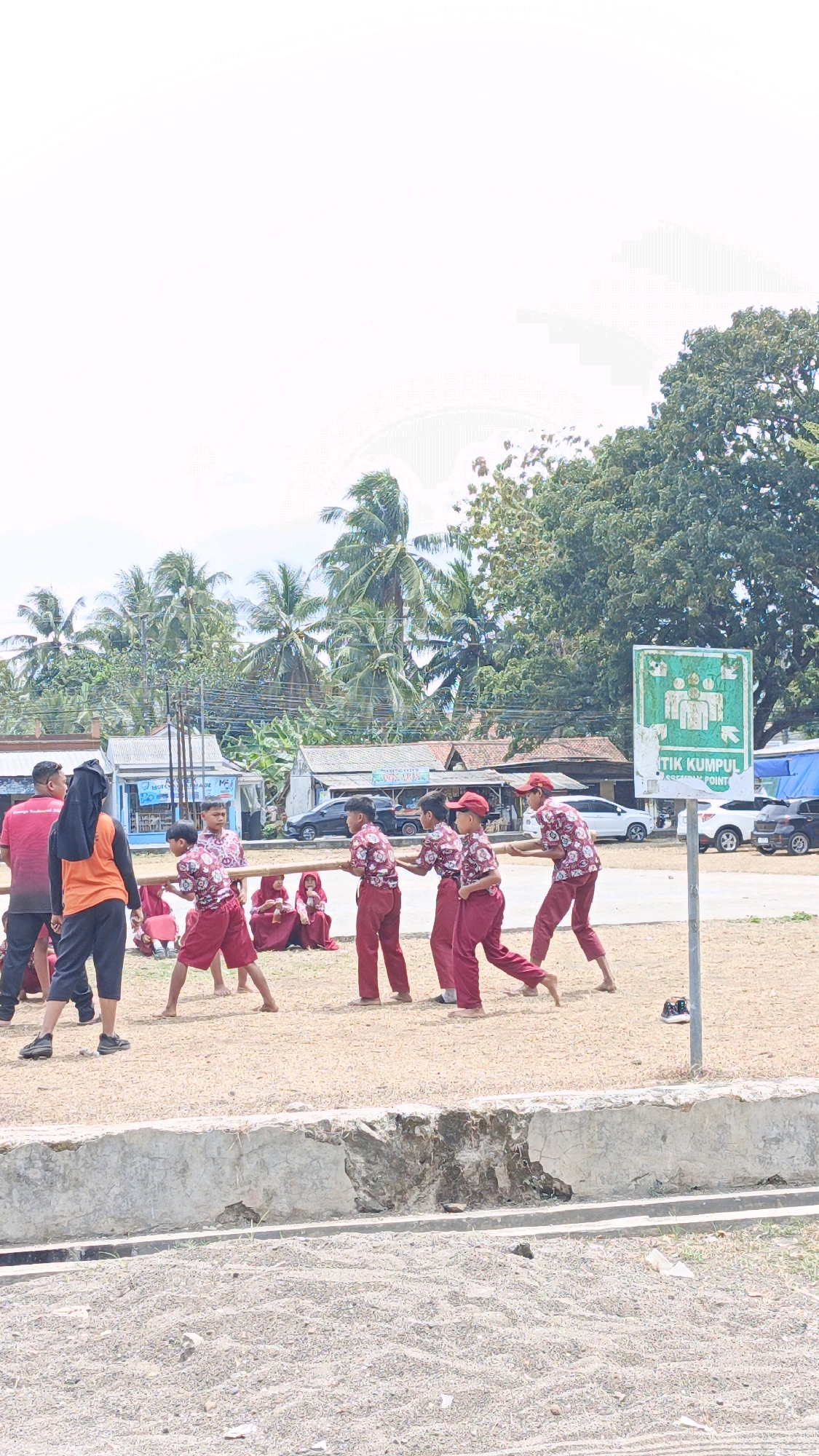 In Sukaresik, Indonesia, a group of schoolchildren in maroon uniforms and red caps are engaged in an outdoor activity. Several children are holding a long wooden pole horizontally, appearing to be in the midst of a game or competition. One child is pulling a rope attached to the pole, suggesting a tug-of-war or a similar team effort. Other children are observing from the sidelines, some sitting on the ground. A few adults are also present, observing the children. The background features a collection of buildings, including shops with signage, palm trees, and a large tree. A green sign indicating a "TIK KUMPUL" (meeting point) is visible on the right side of the image, along with a street with parked cars. The ground is covered in sand and gravel, with a concrete barrier in the foreground. The overall scene depicts a lively event taking place in an open area, likely a schoolyard or public space in Sukaresik.