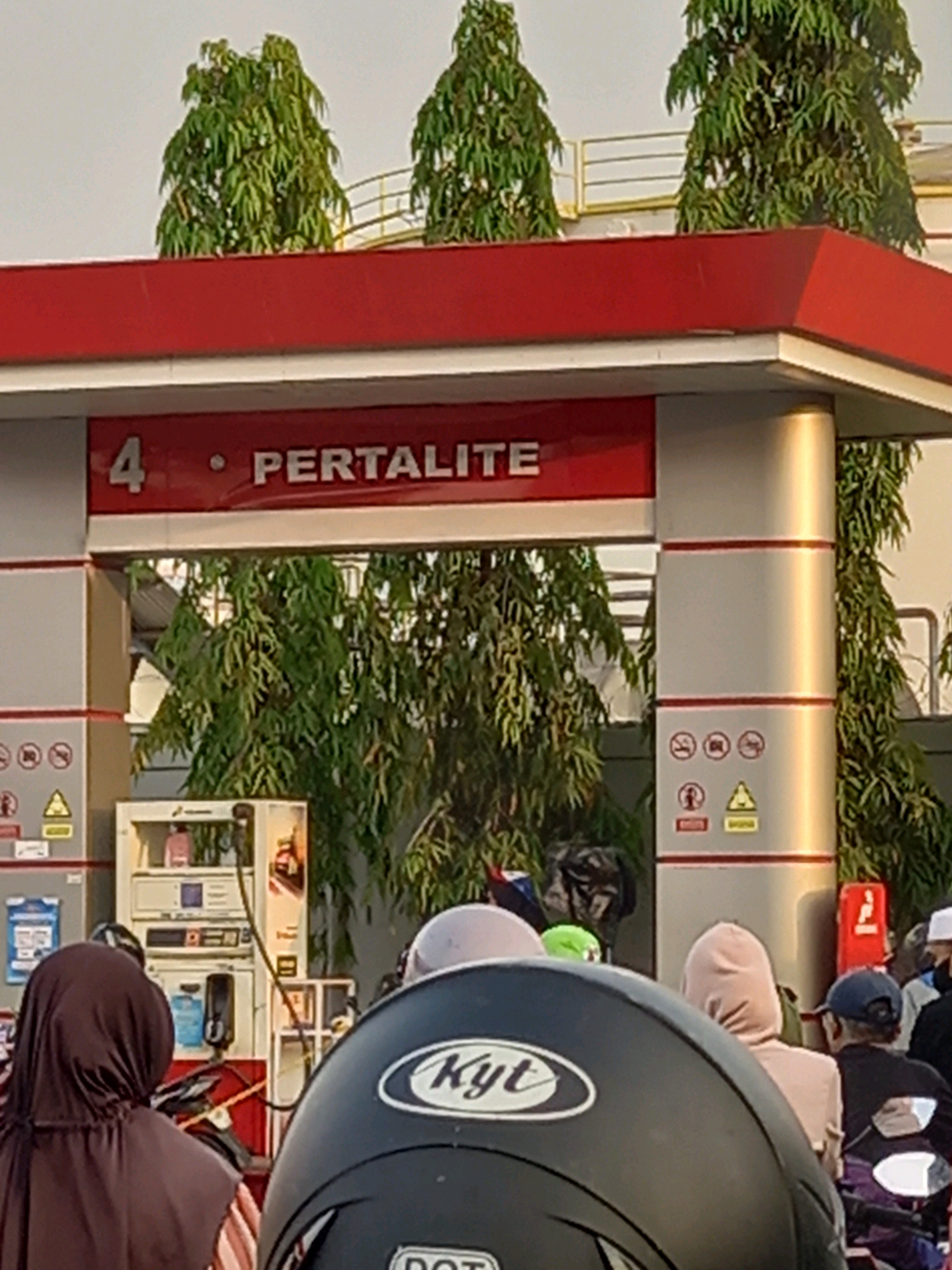 The image captures a scene at a Pertamina gas station, likely in Samarinda, Indonesia, indicated by the "Pertalite" signage. The foreground is dominated by the back of a black motorcycle helmet with "Kyt" branding. Behind it, several people are visible, many on motorcycles, suggesting they are customers.
In the midground, a Pertamina gas pump is present, with various symbols and indicators on its side panels. The pump is white with red accents. Behind the pump, lush green trees with feathery leaves provide a natural backdrop. The gas station canopy is painted red, with "4" and "Pertalite" written in white lettering.
In the background, glimpses of a yellow railing and a structure that might be part of the gas station's infrastructure are visible, partially obscured by the trees. The sky is a pale, overcast color, suggesting it could be daytime. The overall impression is a typical day at a busy gas station, with a focus on the fuel being sold and the people utilizing the services.