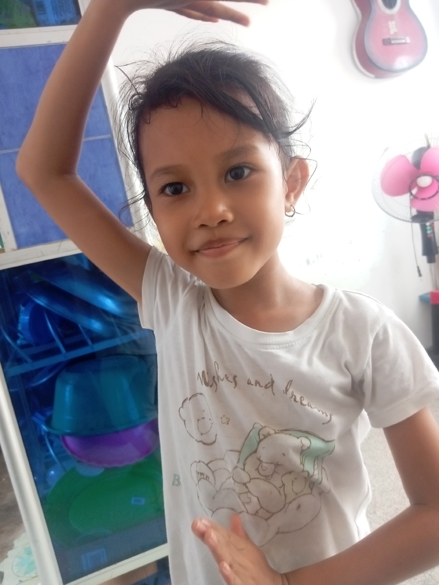 A young girl with dark hair and brown eyes is standing in front of a blue tiled wall and a white wall. She is wearing a white t-shirt with a cartoon bear print and the words "Wishes and Dreams" printed on it. Her right arm is raised above her head. In the background, there is a pink electric fan and a decorative guitar on the wall. To the left of the girl, there is a cabinet with blue and green bowls inside. The image appears to be taken indoors, likely in a residential setting in Batam, Indonesia.