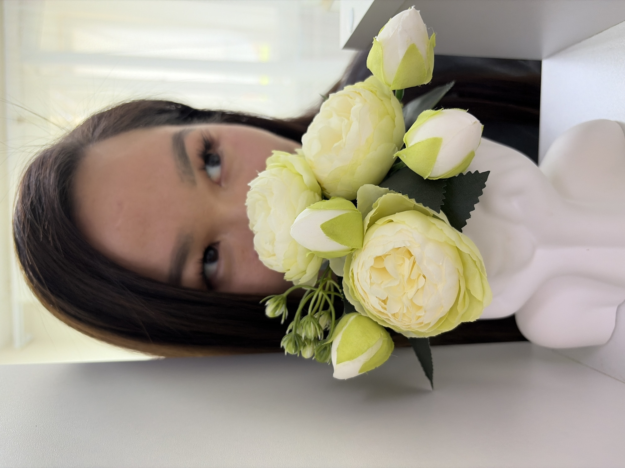 A close-up shot features a woman partially visible behind a bouquet of flowers and a white object. The woman has dark brown hair, visible eyebrows, and eyes that are the focal point. The bouquet is comprised of white and yellow flowers with green leaves. The white object behind the flowers appears to be a vase or decorative piece. The background includes hints of a window and what seems to be a shelf or table the objects are placed on. The image appears to be taken indoors. The location in the city of Unknown, Unknown, is not discernible from the provided image.