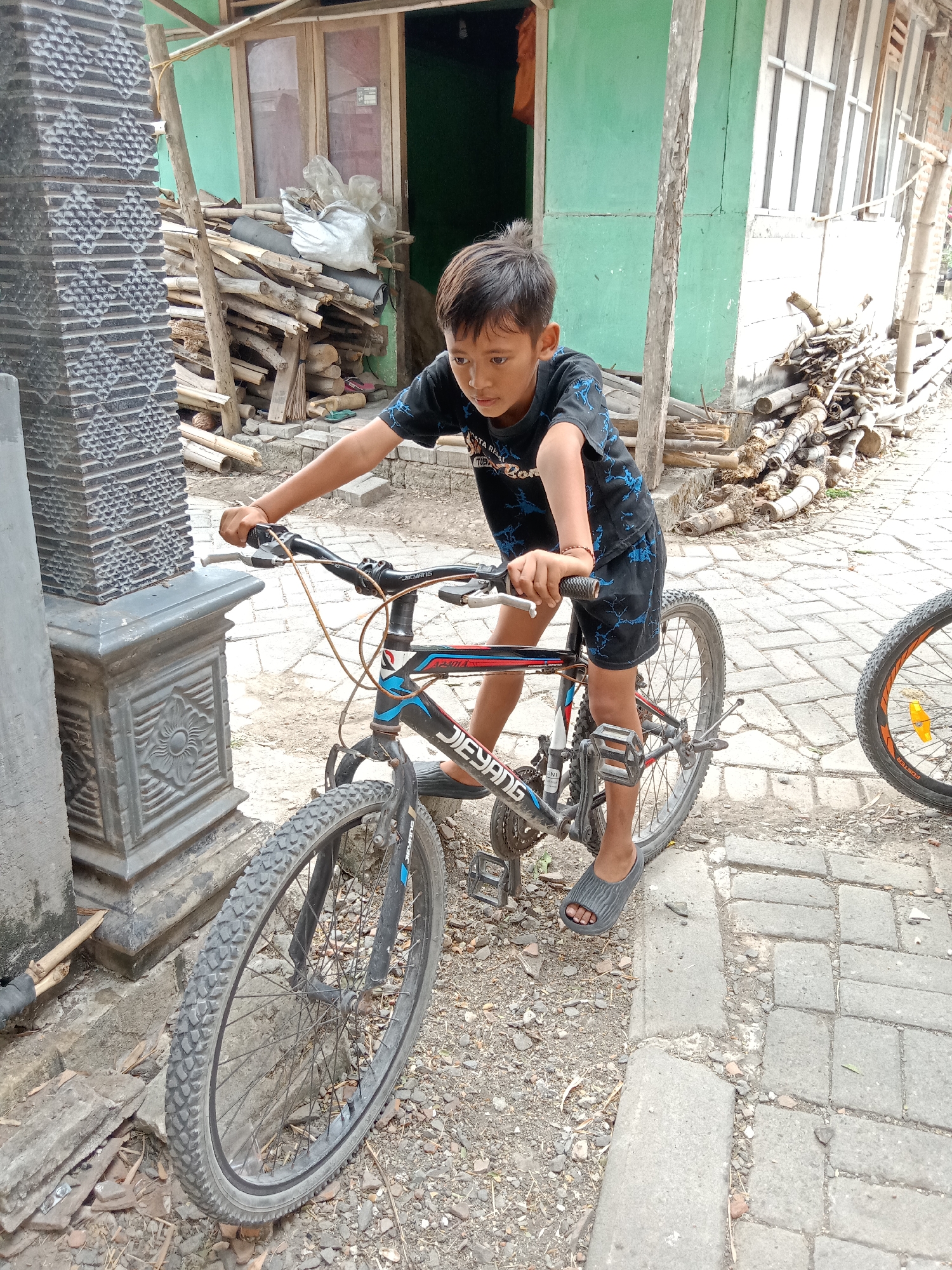 In Jumput, Indonesia, a young boy is pictured on a bicycle. The boy, wearing a black t-shirt with a blue design and black shorts, is holding the handlebars of a black and blue mountain bike. He is wearing grey flip-flops. The bicycle has a silver frame, black tires, and a visible gear system. The boy appears to be focused on something out of frame to his left.
The environment suggests a rural or semi-urban setting. To the left of the boy is a grey textured pillar. Behind the boy, a building with a green wall and wooden-framed windows is visible. Piles of cut wood and bamboo are stacked near the building. The ground is paved with grey bricks and covered with gravel and dirt. Another bicycle wheel, with an orange rim and black tire, is partially visible on the right side of the frame. The lighting indicates it is daytime.