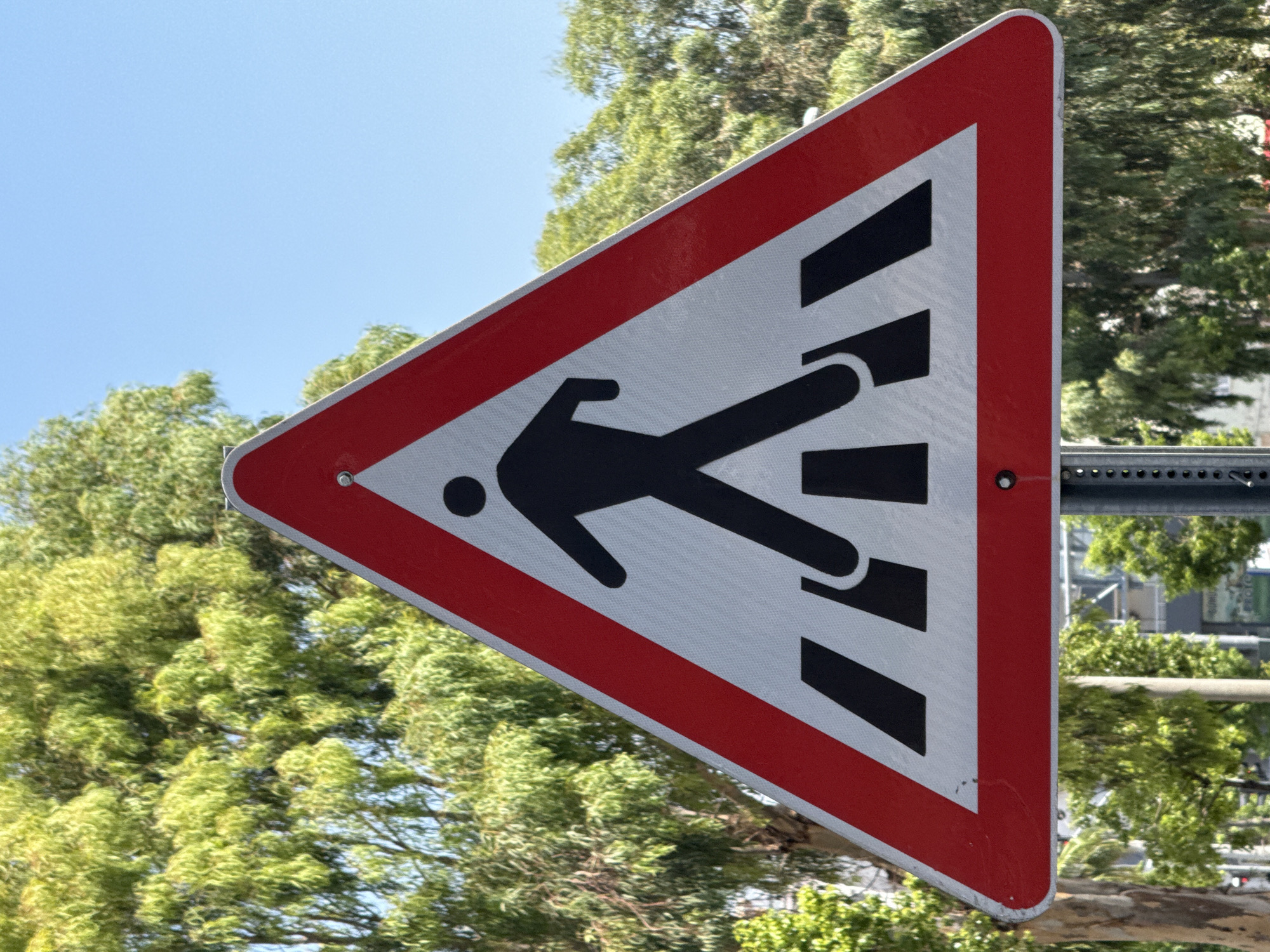 The image shows a pedestrian crossing warning sign, commonly known as a zebra crossing sign, positioned outdoors in Silifke, Turkey. The sign is triangular with a red border and a white interior, featuring a black silhouette of a person walking on a zebra crossing. Behind the sign, lush green trees are visible against a clear blue sky. In the background, some buildings and a street are discernible. The sign is mounted on a metal pole. The overall scene suggests an urban or semi-urban environment where pedestrian safety is a consideration.