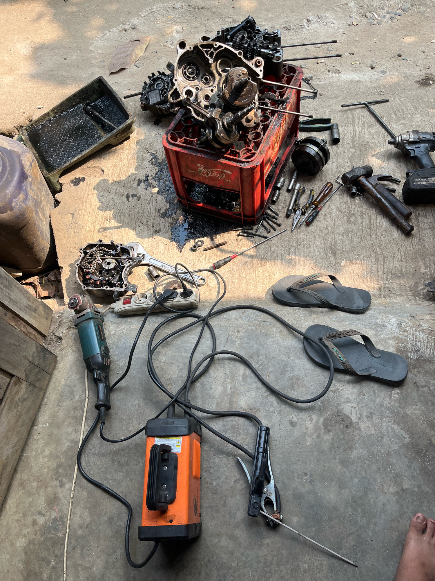 The image depicts an outdoor workshop area, likely on a concrete ground, in Kebumen, Indonesia. The scene is cluttered with various tools and disassembled motorcycle engine parts. In the center of the frame, a red Coca-Cola crate holds a partially disassembled engine, revealing gears and crankshaft components. To the left, a black oil pan is filled with dark liquid, and a large, partially empty plastic container sits nearby. A green angle grinder, an orange welding machine with cables and a clamp, and a power strip with connected plugs are also present. Scattered around the engine parts are wrenches, pliers, screwdrivers, a hammer, a socket wrench, and a cordless drill. Two pairs of dark flip-flops are placed on the ground, and a human foot is visible in the bottom right corner of the image. The ground is a mix of concrete and dirt, with shadows indicating sunlight. The overall impression is of a makeshift repair or maintenance station.