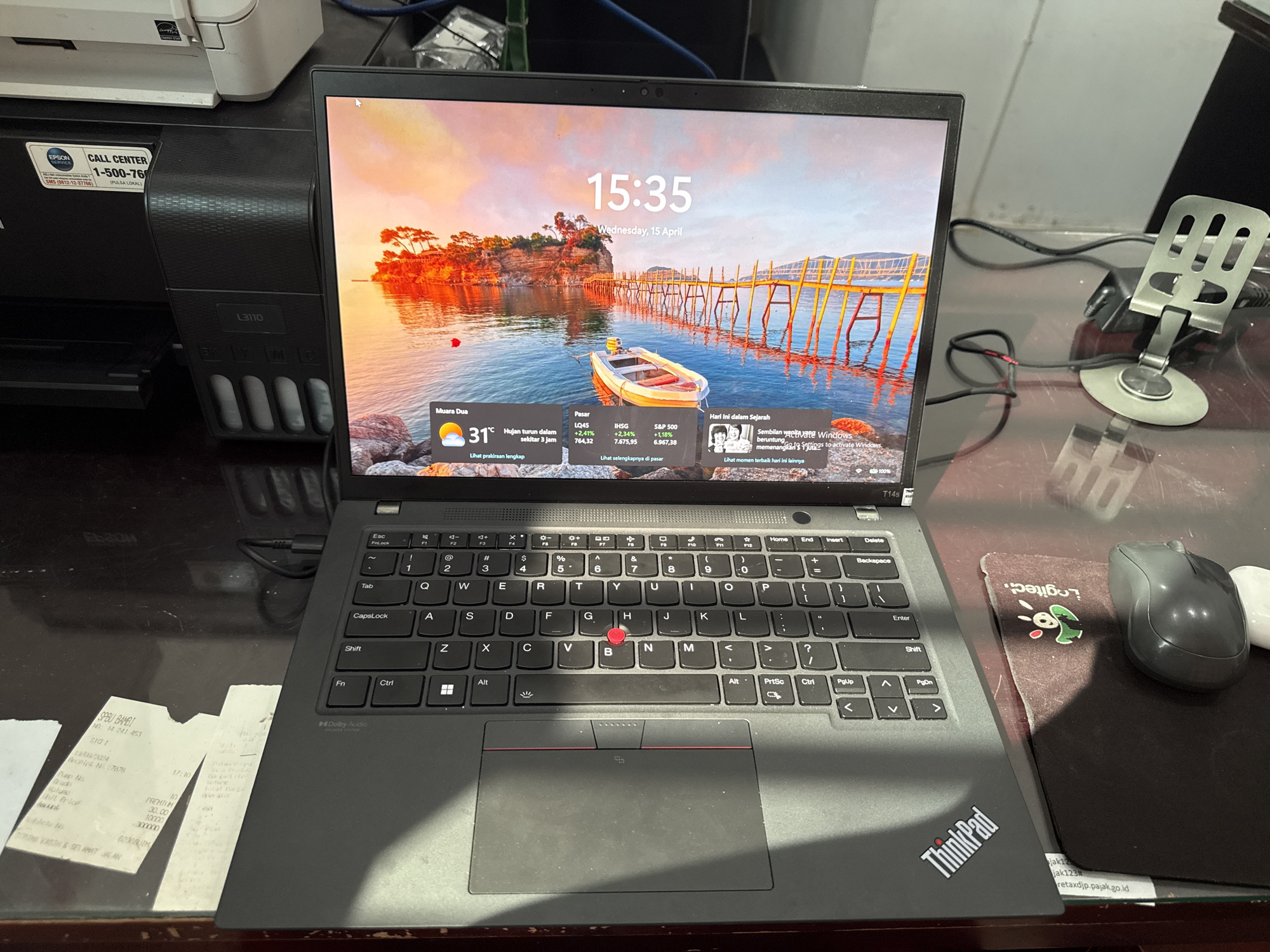 The image depicts a Lenovo ThinkPad T14s laptop displaying a screen with the time 15:35, Wednesday, April 15, along with weather and stock market information. A fuel purchase receipt is to the left of the laptop, and a Logitech computer mouse is to the right. An Epson printer is visible in the background. The date on the fuel receipt is 13/08/2X224.
Based on the weather information (Muara Dua) and the location on the fuel receipt, the location is likely Lhokseumawe, Indonesia.