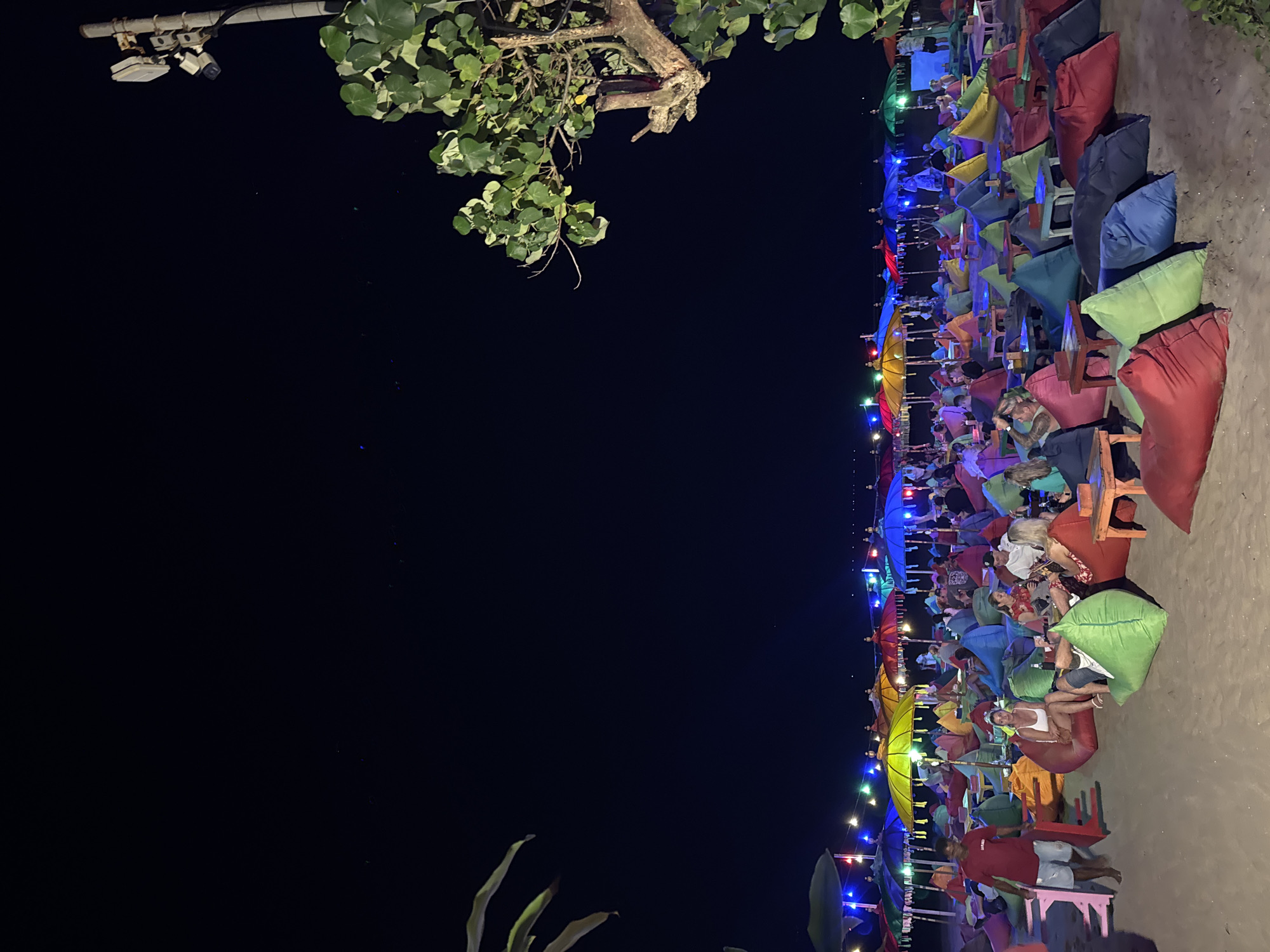 A crowded beach scene at night in Kuta, Indonesia features numerous people seated on colorful beanbags. They are spread out across the sand under a canopy of brightly colored umbrellas and string lights, creating a festive atmosphere. Small tables are interspersed among the seating areas. In the foreground, a man in a red shirt walks towards the left, while groups of people are engaged in conversation and relaxation. To the right, a large tree with green leaves extends into the frame, and a utility pole with surveillance equipment is visible against the dark night sky. The beach is sandy, and the overall impression is that of a lively nocturnal gathering.