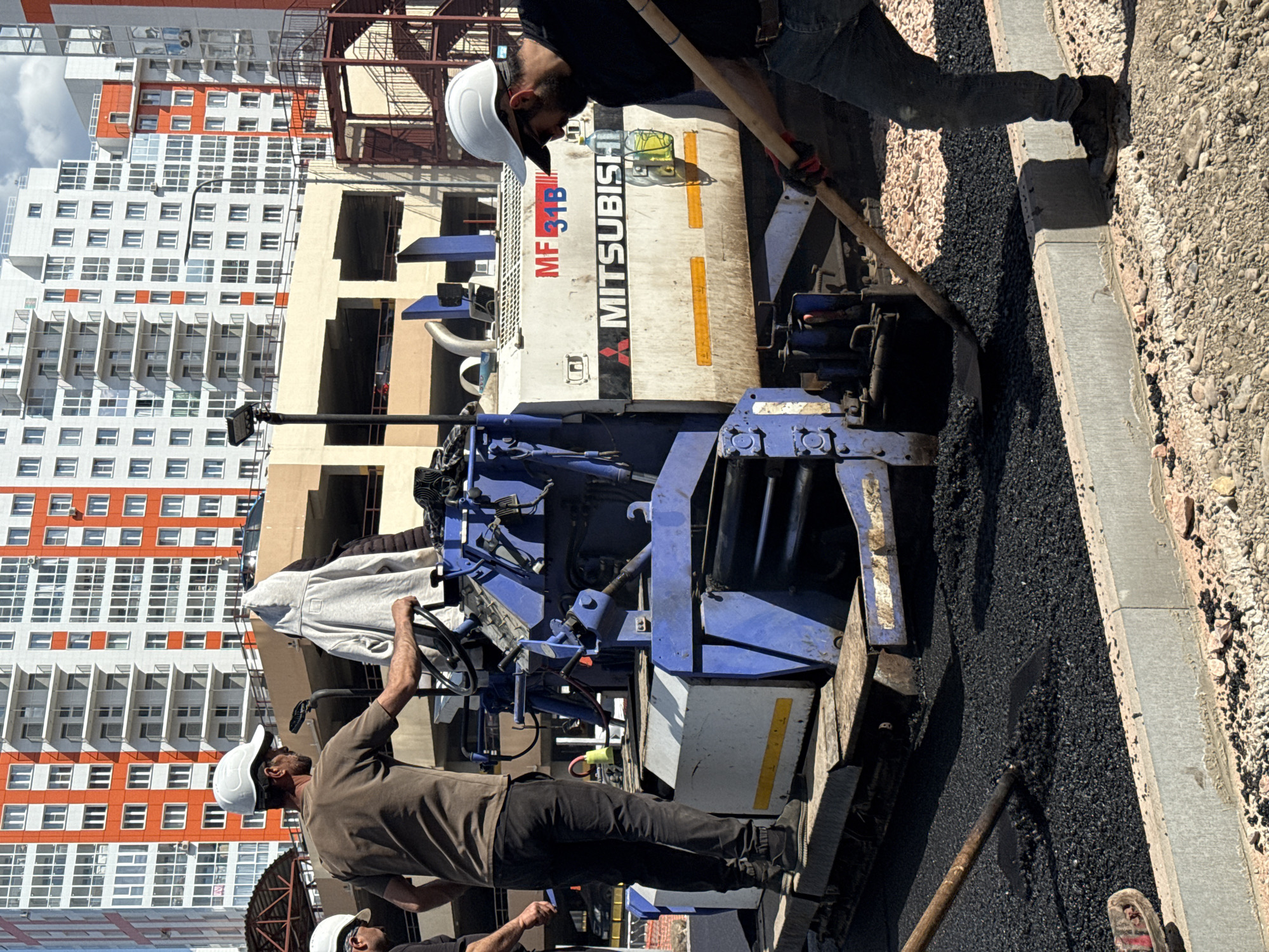 Two construction workers are using a Mitsubishi MF 31B asphalt paver to pave a street in Krasnoyarsk, Russia. One worker is operating the paver. The other worker is using a shovel to smooth the asphalt. A large apartment building with white and orange accents is in the background.