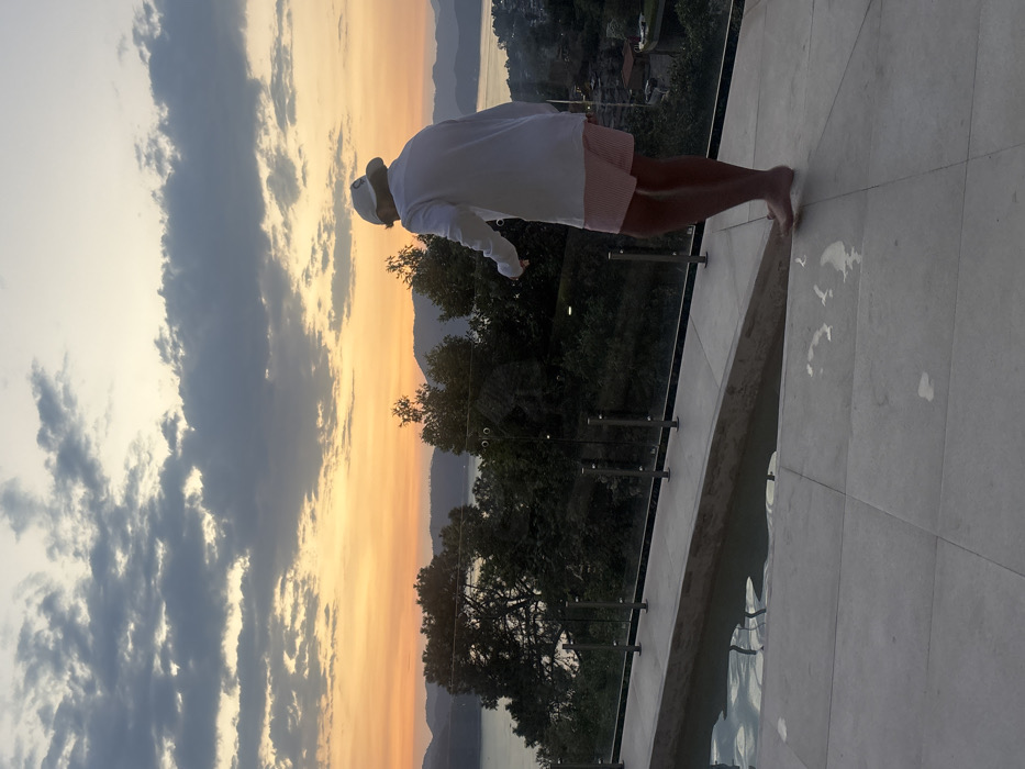 An elevated view from a poolside terrace in Valle de Bravo, Mexico, depicts a person observed from behind during sunset. The sky displays an orange and yellow gradient at the horizon, transitioning to light blue and scattered gray clouds overhead. Multiple layers of dark hills and mountains are visible in the distance, framing a large body of water, likely the lake characteristic of Valle de Bravo. Dense green trees occupy the midground slopes, partially obscuring a distant urban area with scattered structures visible on the right.
In the foreground, a light-colored paved terrace extends across the frame. To the left, an infinity-edge pool with water is partially visible. A transparent glass railing with several stainless steel posts runs along the edge of the terrace, overlooking the landscape.
A single individual, a human, stands on the paved terrace, facing away from the camera and looking towards the distant view. The person is barefoot, with distinct wet footprints visible on the light-colored tiles. They are dressed in a white long-sleeved top or light jacket, pink and white striped shorts, and a white baseball cap featuring a dark emblem on the rear. The individual's right leg is slightly lifted, indicating a motion of walking or shifting stance. No other individuals are present within the frame.