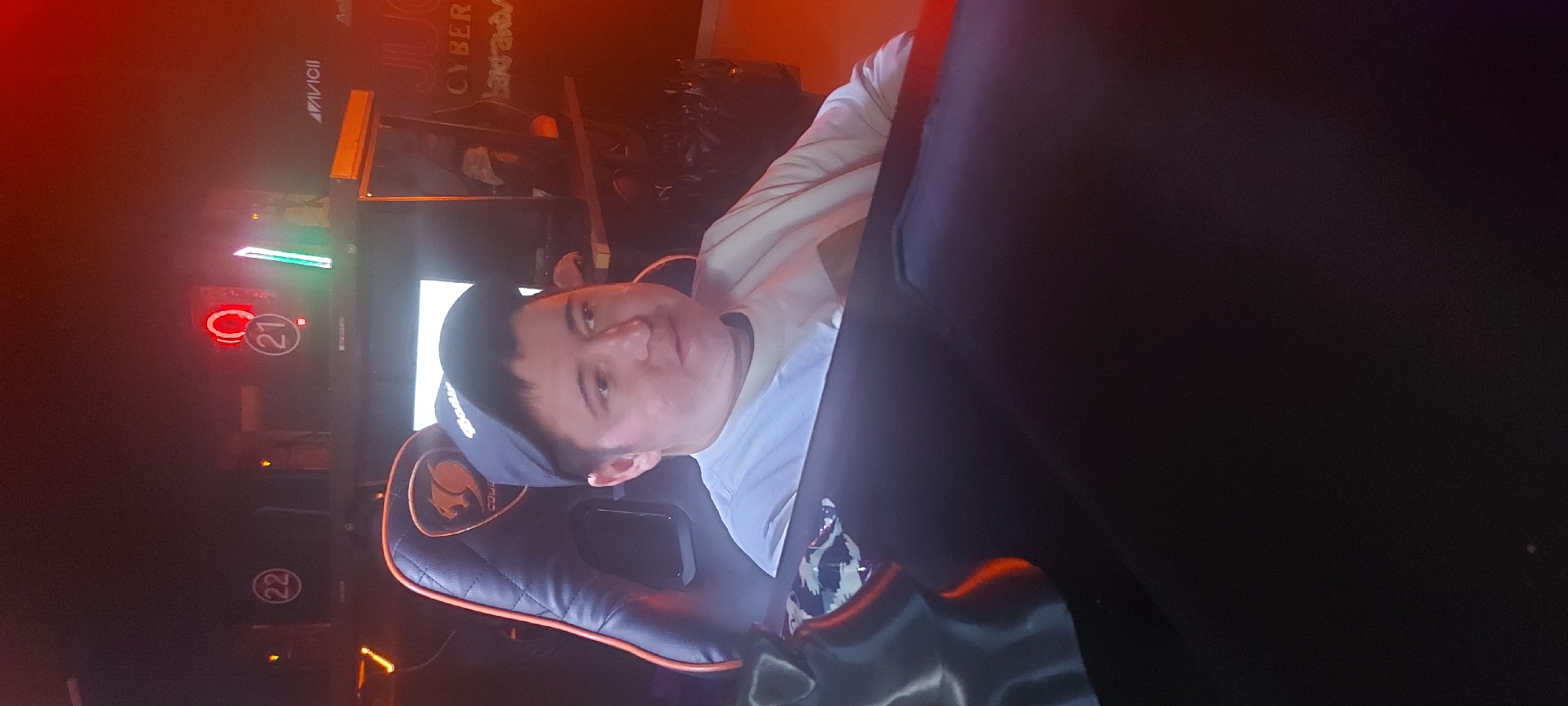 The image, oriented vertically, depicts an indoor setting, likely a gaming cafe or computer club in Tashkent, Uzbekistan. A young man is the primary subject, seen from the chest up, looking directly at the camera. He is sitting in a black and orange/brown gaming chair, identifiable by a visible Cougar brand logo on its headrest. He is wearing a light-colored t-shirt and a dark baseball cap.
The background is dimly lit, dominated by artificial light sources. To the man's left, a bright, rectangular white screen is visible, likely a monitor. Further to the left, a vertical rack or computer station is illuminated with various colored LED lights, including green and red. Prominently featured on this station are two circular, glowing red outlines containing white numbers: "21" at the top and "22" below it, indicating station numbers.
In the upper background, reflected on what appears to be a mirror or glass surface, partial text is visible. "AVICII" is clearly legible, and the word "CYBER" is also present, suggesting a business name like "CYBER SPORTS" or "CYBER CAFE." The overall atmosphere is dark with glowing electronic elements. The man has a neutral to slightly pleasant expression, seemingly pausing for the photo.