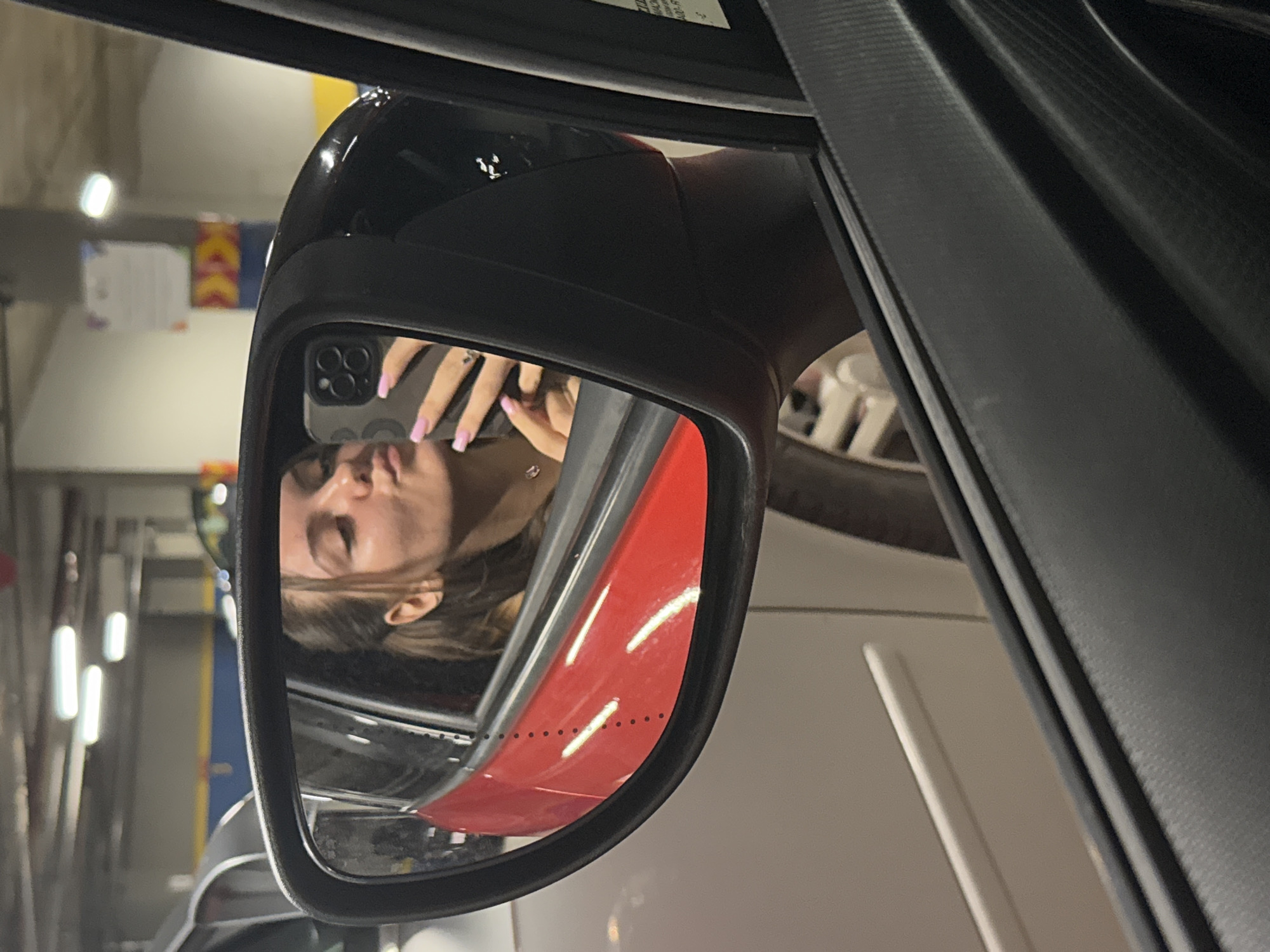 The image shows a reflection in a car's side mirror. A person, likely a young woman, is visible taking a selfie with a smartphone. The reflection includes a partial view of a red and white car next to the vehicle from which the photo is taken. The setting appears to be an indoor parking garage with fluorescent lighting. It is not possible to determine the location is in Sofia, Bulgaria from the image.