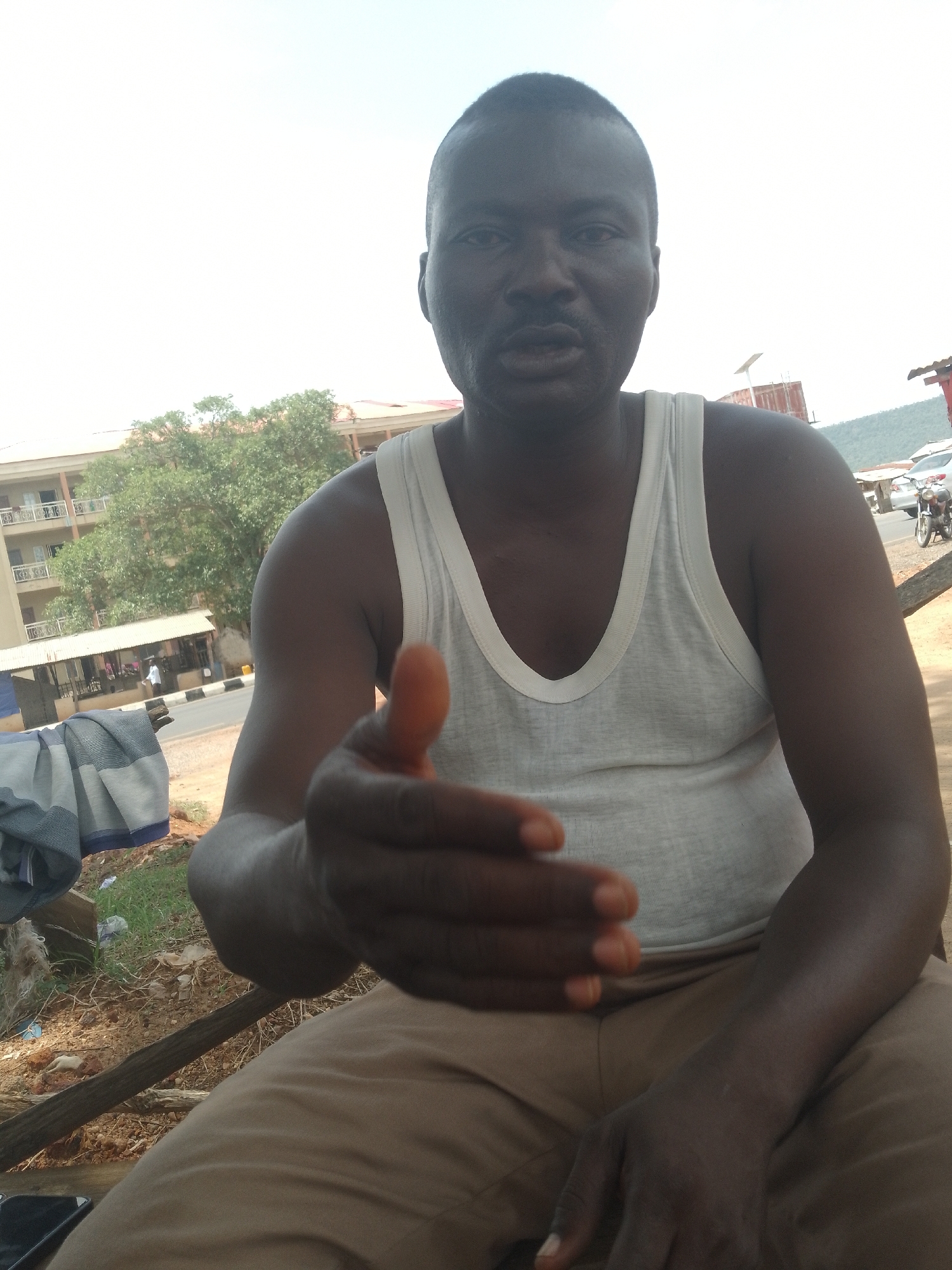 A dark-skinned man in a white tank top and khaki pants sits outdoors in Adankolo, Nigeria, extending his hand toward the camera. Behind him, across a road, is a multi-story building, trees, and a small covered structure. A motorcycle and parked cars are also visible. A piece of fabric is draped to his left. The area around him appears to be a roadside with some dirt and sparse vegetation.