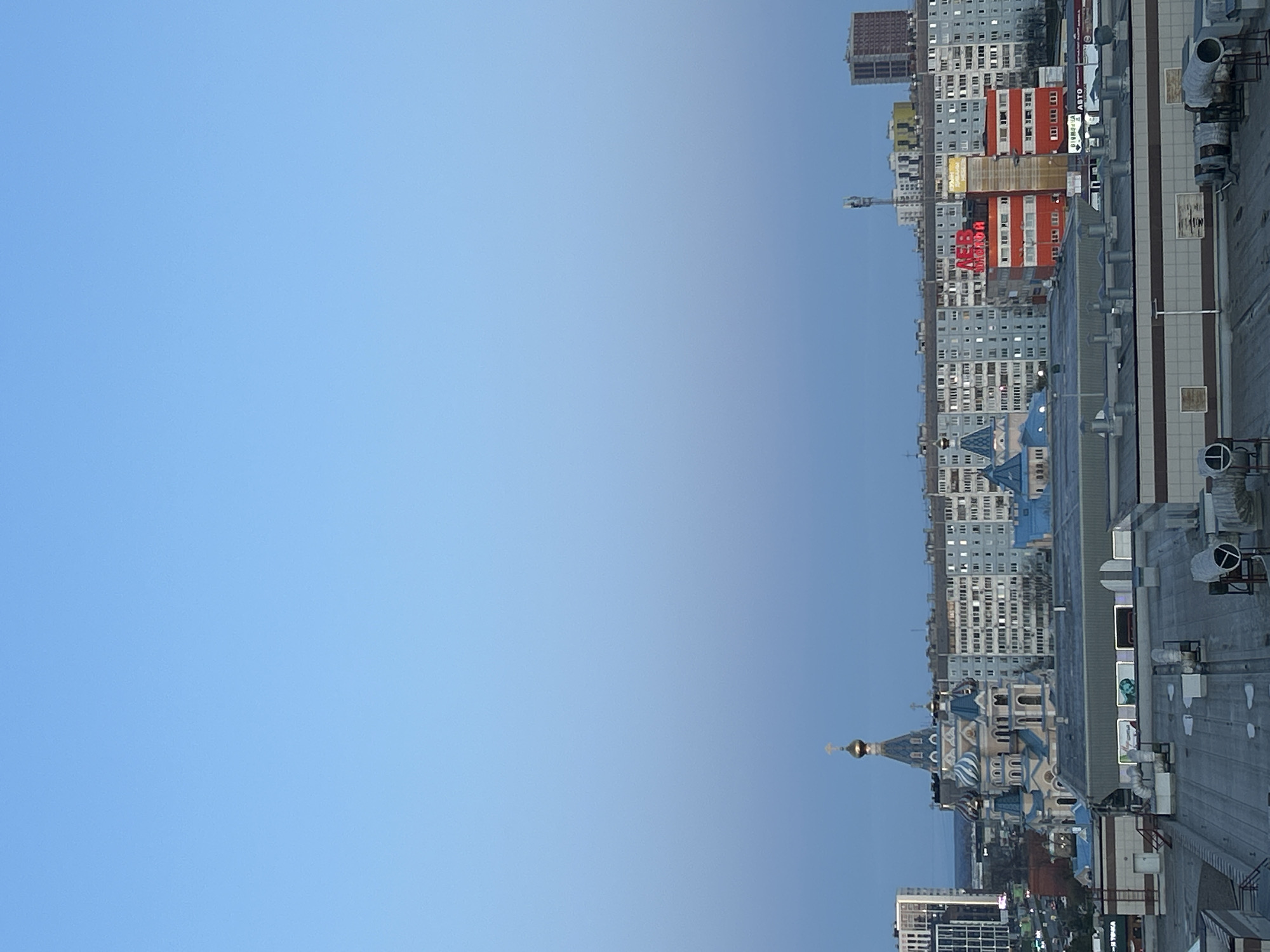 This image, captured from an elevated perspective, offers a panoramic view of an urban landscape in Izhevsk, Russia, under a clear, bright blue sky. The original image is rotated 90 degrees clockwise, but when viewed correctly, the horizon line is set high, dedicating a large portion of the frame to the expansive sky.
In the immediate foreground on the right, a flat industrial rooftop is visible, equipped with various ventilation shafts, pipes, and air conditioning units, suggesting a commercial or large residential building. Beyond this rooftop, the city unfolds with a dense collection of buildings.
A distinctive Orthodox church stands prominently in the mid-ground, characterized by its ornate blue and white patterned facade, multiple domes, and golden onion-shaped cupolas topped with crosses. Surrounding the church are numerous multi-story residential and commercial buildings, predominantly blocky in design with repetitive window structures. Notable among these is a building with a red and white horizontally striped or paneled facade, and another taller, darker, possibly brick-clad building further in the background.
The sky, occupying the upper and left portions of the image, is a uniform light blue, completely free of clouds, indicating fair weather during daylight hours, likely morning or late afternoon given the soft, even illumination.
Visible text includes a prominent red sign on the red and white building that reads "ДВЕНАДЦАТЬ" (DVENADTSAT', meaning "Twelve") in Cyrillic. Closer to the foreground rooftop, a white sign with black text displays "АВТО" (AVTO, meaning "Auto"), also in Cyrillic. No specific human activity or event is captured, presenting a static, observational cityscape. The overall impression is one of a vast, settled urban environment.