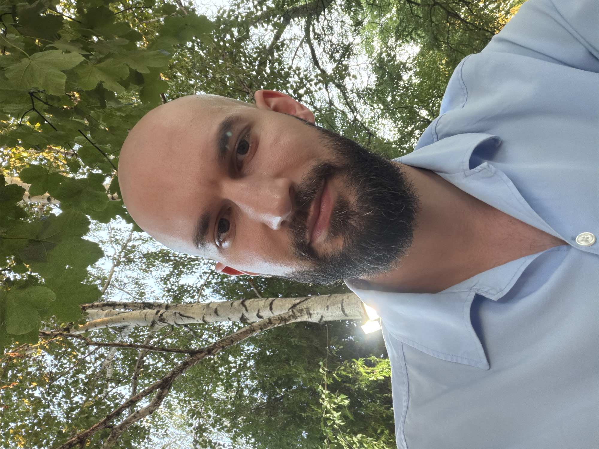 The image is a close-up, low-angle selfie of a bald man with a beard, wearing a light blue collared shirt. He is positioned in the foreground, with trees and foliage forming the background. The lighting suggests it is daytime, possibly late afternoon, with sunlight filtering through the leaves. The setting appears to be an outdoor environment, consistent with a park or garden. There is no visible indication of the specific location being Mursalevo, Bulgaria.