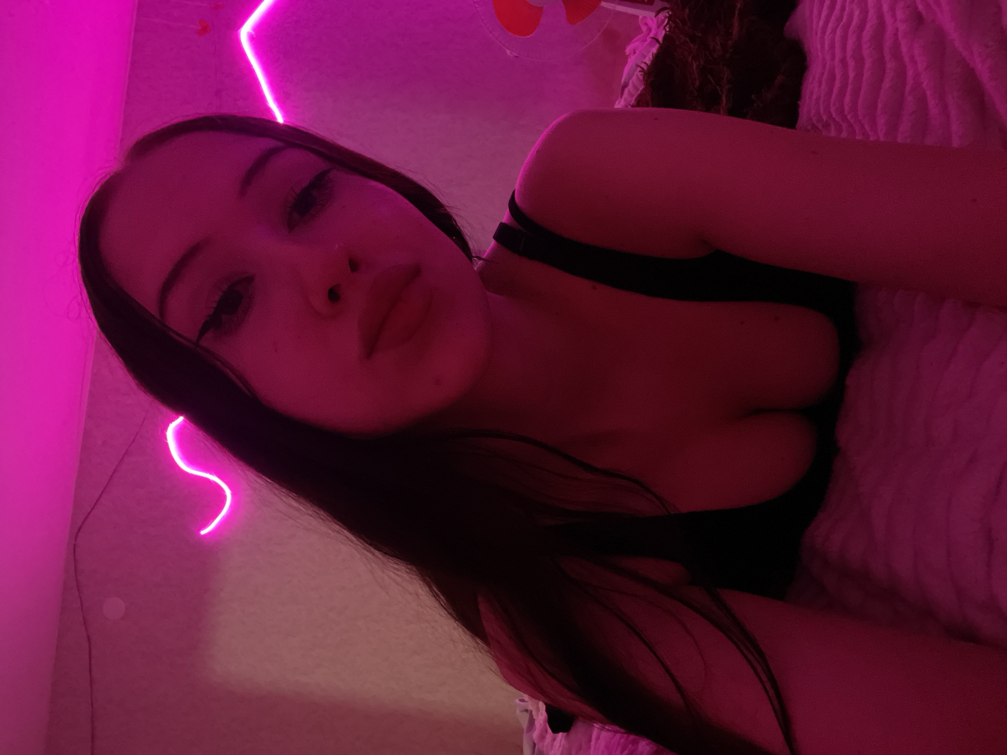 A young woman with dark hair, wearing a black top, is lying on a bed. She is positioned in the foreground of the image, looking towards the camera. The room is illuminated by pink neon lights, which cast a vibrant glow on the surroundings. The bed is covered with a textured pink blanket. In the background, more pink neon lights are visible on the wall, forming abstract shapes. There is also a glimpse of a decorative item, possibly a fan, to the right of the woman. The overall impression is a dimly lit, atmospheric bedroom setting. The image does not contain any specific geographical markers that would indicate a location in Votkinsk, Russia.
