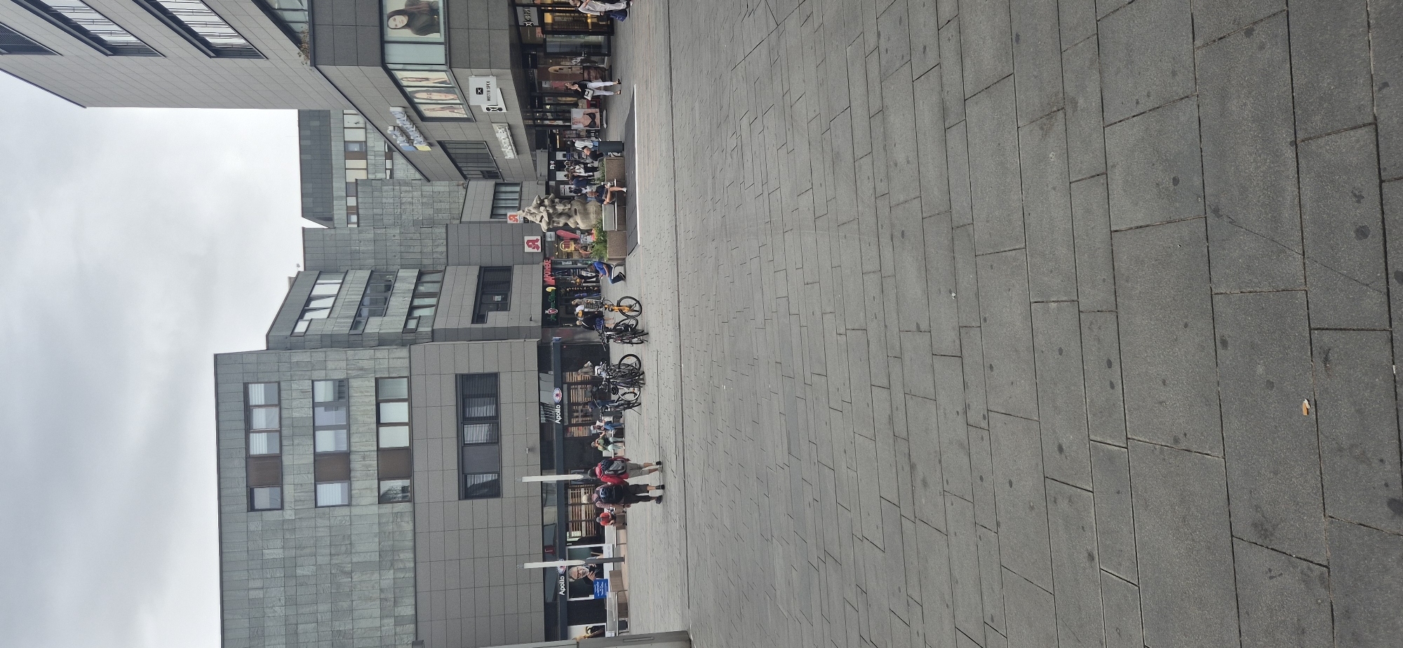 The image depicts a daytime urban scene in Mainz, Germany, likely in a pedestrianized commercial area. In the foreground, a wide expanse of gray, rectangular paving stones stretches into the distance. Several bicycles are parked on the pavement. In the mid-ground, people are walking and sitting. On the left, two individuals are walking away from the camera. Beyond them, shops with large glass windows are visible, including "Apollo" which appears to be an optician. Further back, a fountain sculpture stands with people gathered around it. The background is dominated by modern, multi-story buildings with grey facades and numerous windows. The sky is overcast.