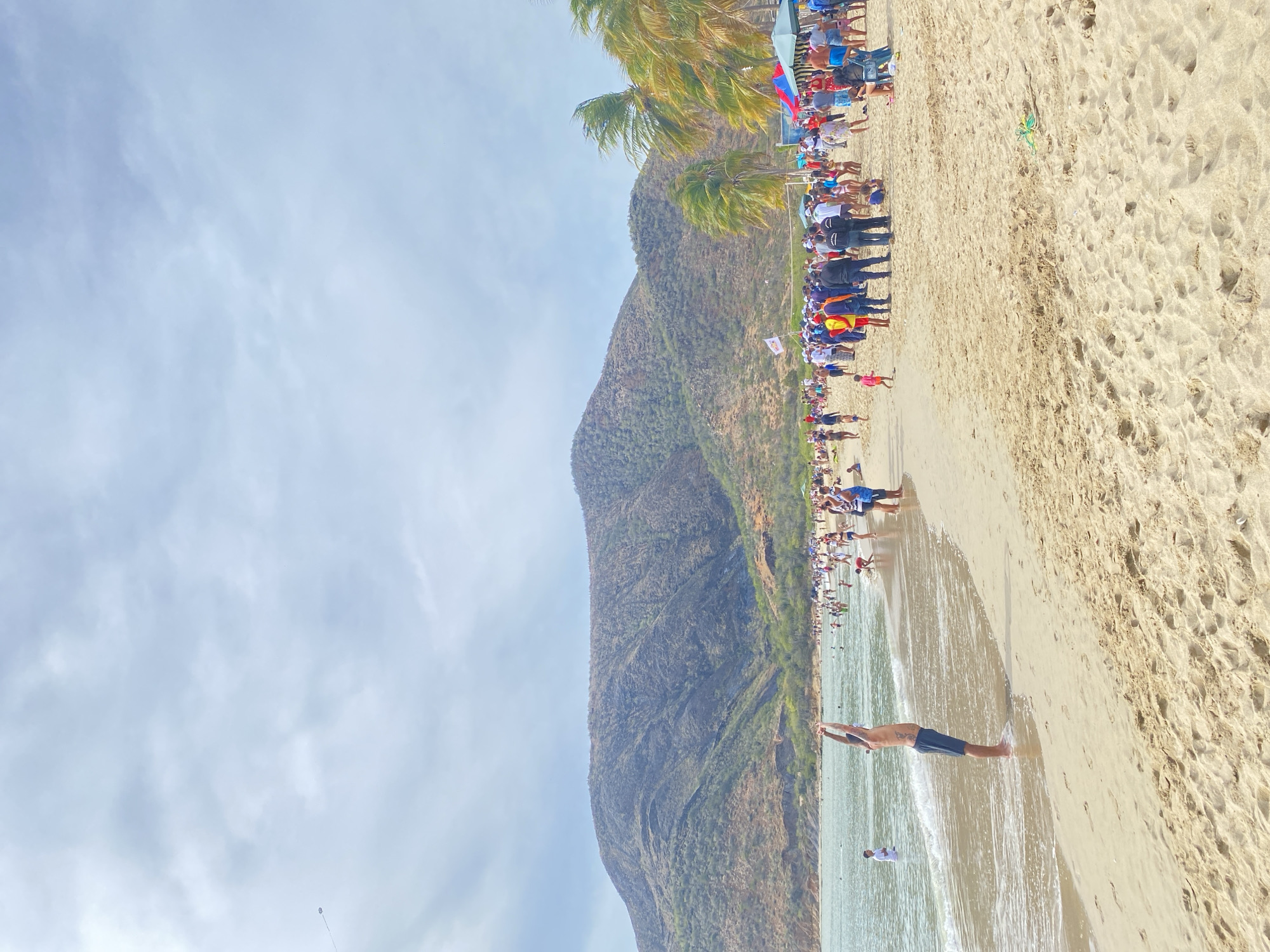 The image depicts a crowded beach in Juan Griego, Venezuela, adjacent to a mountain. The beach is filled with people, many gathered near the shoreline, and some in the water. The beach is sandy with visible footprints and tidal lines. One man is diving into the water. The mountain rises steeply behind the beach and is covered in vegetation. Palm trees are visible along the edge of the beach where people are gathered. The sky is cloudy, with sunlight breaking through. Some people have flags or are wearing matching outfits, suggesting a group event.