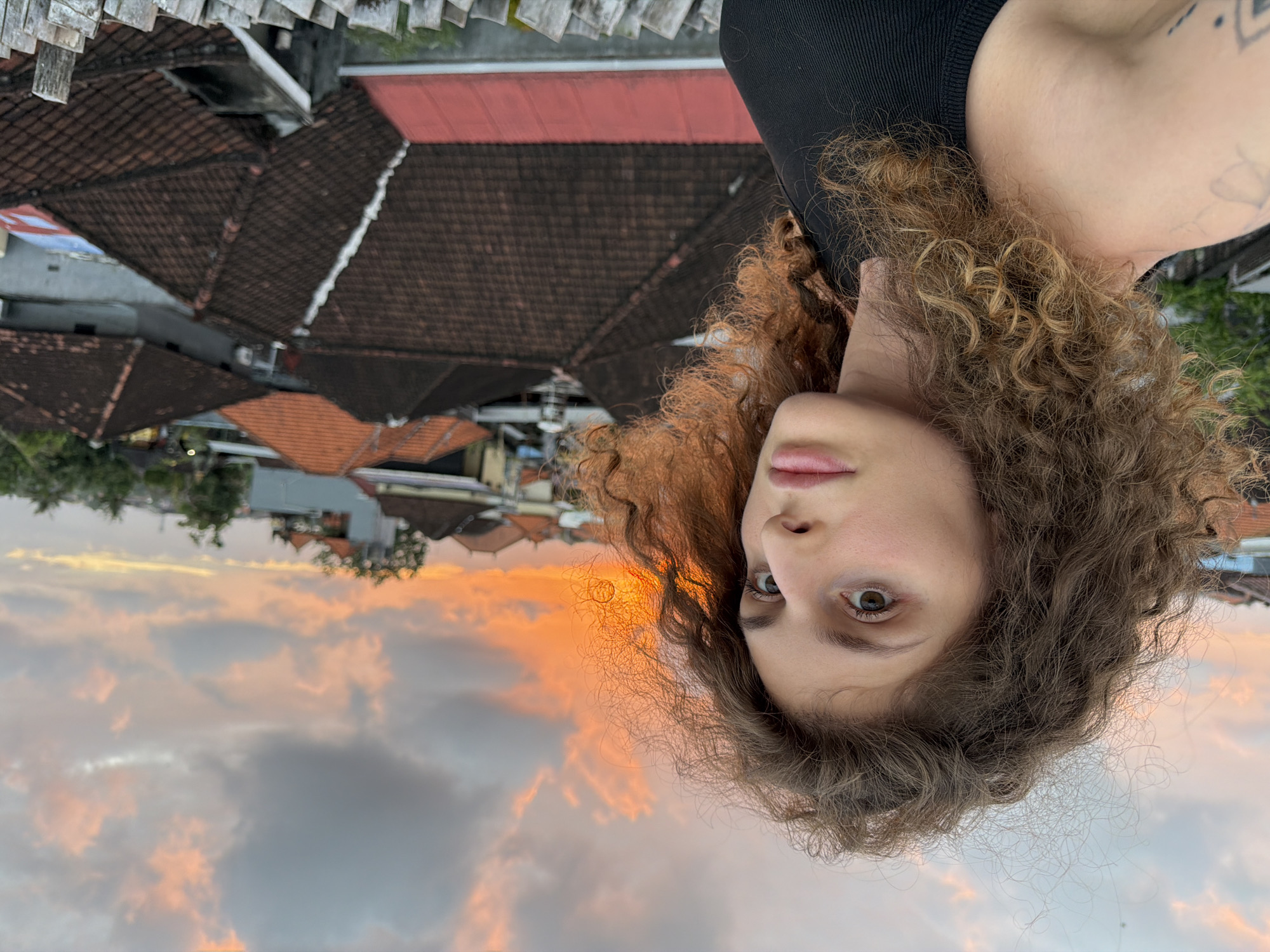 A young woman with curly brown hair is taking a selfie outdoors. She is wearing a black top. Her left shoulder and arm are visible, with a tattoo on her upper arm. The background shows a tiled roof landscape, typical of North Kuta, Indonesia, with the sky at sunset in the distance. The sky is filled with clouds in shades of orange, pink, and gray, indicating a dramatic sunset. The scene appears to be on a rooftop or balcony, suggesting an urban or suburban setting with residential buildings. The woman's expression is neutral, with her eyes looking directly at the camera. The overall mood is relaxed and natural. No visible text is present in the image.