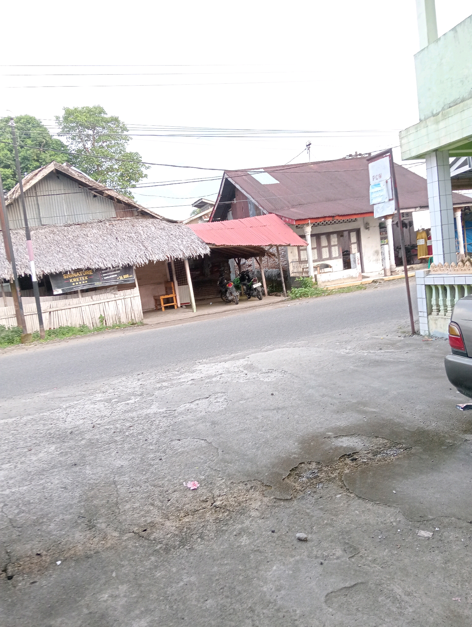 The image depicts a street scene in Campago, Indonesia. On the left side of the road, a building with a thatched roof and a corrugated metal upper section is visible. A sign with "SIGNATURE KRETEK" is hanging from the front of this building. Adjacent to it, there is a covered area with a red roof where two motorcycles are parked. Further down the street, a building with a dark red tiled roof and white columns is seen. A sign with "FCW" is mounted on a pole in front of this building. A light green building with white decorative pillars is on the far right of the image. The road surface is asphalt and shows signs of wear and tear, with visible cracks and patches. The sky is overcast. There are no people clearly visible in the image.