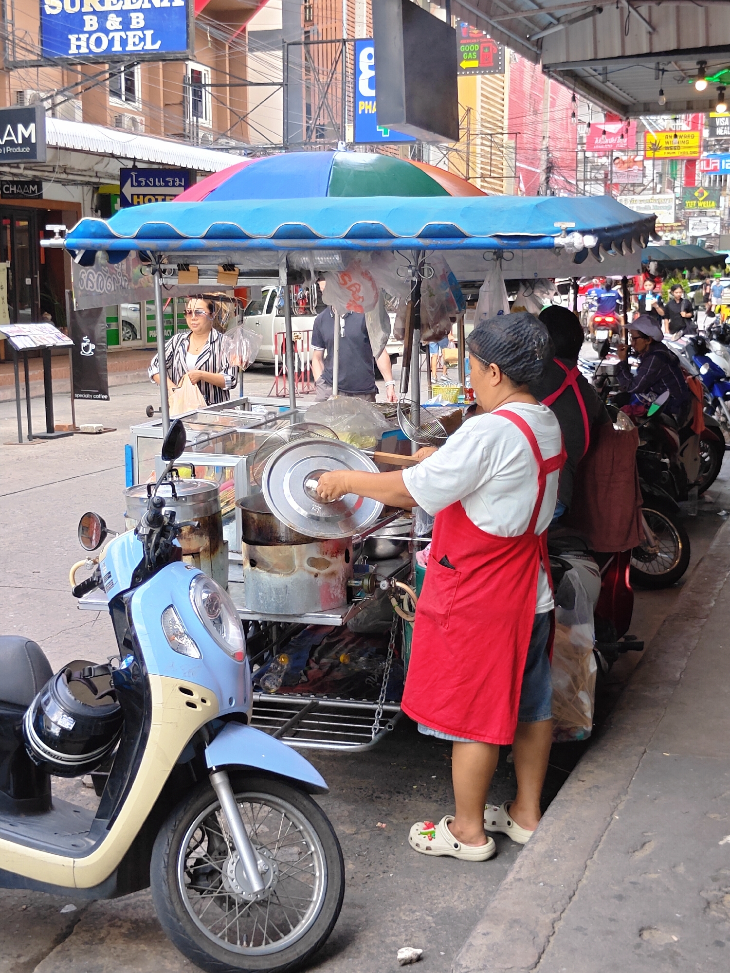 A street scene in Pattaya City, Thailand, shows a food cart with a blue and green striped umbrella. The cart is attached to a light blue and cream colored scooter. A woman wearing a red apron and a white t-shirt, with a black hairnet, stands behind the cart, holding a metal utensil. She is looking down at the cart's contents. Another person, wearing a black t-shirt and a red apron over it, is partially visible behind her. A woman in a striped shirt and sunglasses is standing to the left of the cart, holding a plastic bag. In the background, several motorcycles are parked, and there are buildings with signs in Thai and English, typical of a commercial street. The overall scene depicts a vibrant street food vendor operation on a moderately busy street.