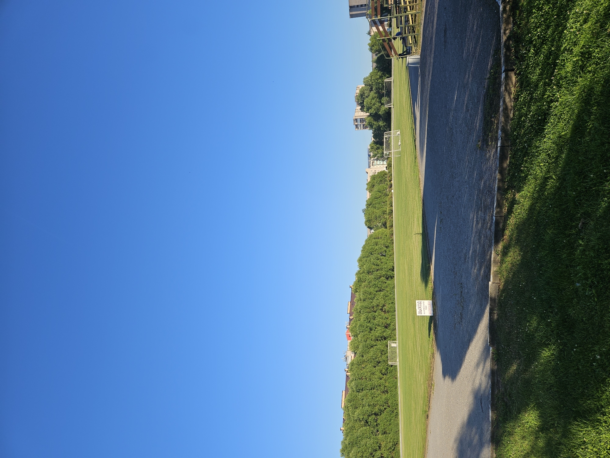 This is a vertical, eye-level shot of a football field in Veliky Novgorod, Russia, on a clear, sunny day. The field is green and well-maintained, with two soccer goals visible in the distance. A row of dense, green trees lines the back of the field, with apartment buildings peeking out above them. In the foreground, a paved area transitions to a grassy area, with shadows cast by an unseen structure. A white sign with Russian text is positioned on the paved area, which appears to be near a walkway. Bleachers, partially visible on the right side, suggest a stadium or public viewing area. The sky is a brilliant, cloudless blue, indicating favorable weather conditions.
