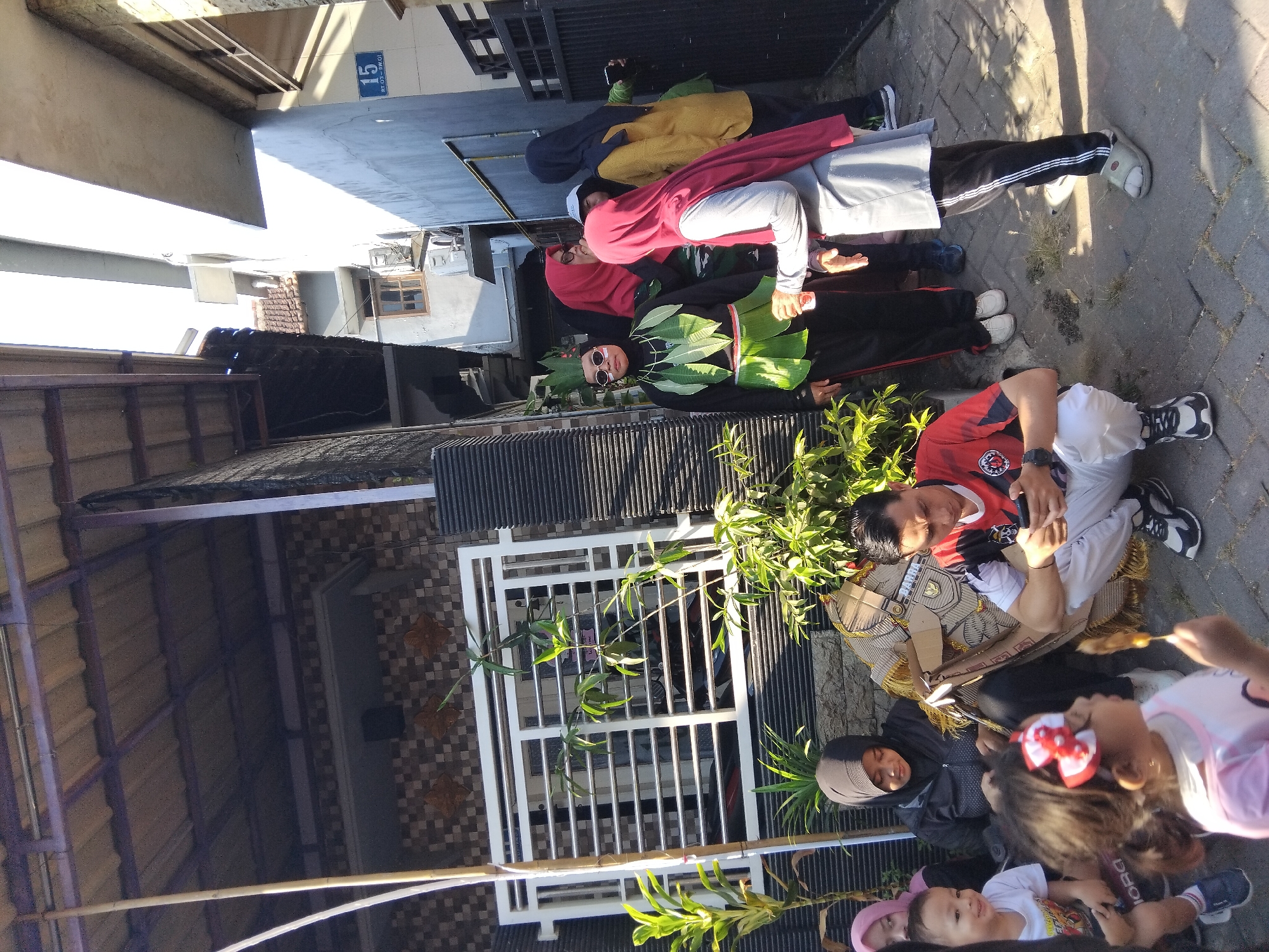 In the city of Kepuh Kiriman, Indonesia, a group of people are gathered outdoors on a paved walkway. A man in a red and black athletic shirt and white pants is squatting, looking at his phone. Next to him, a child in a pink shirt with a red and white bow in her hair is holding a skewer with food. To the left, a woman in a black hijab is sitting and appears to be holding a piece of fabric. Behind them, a young child in a white t-shirt is smiling. Further back, a woman in a black hijab and a pink hijab are visible. In the foreground, a person wearing a costume with green leaves and sunglasses is standing. Other individuals, including a woman in a red hijab and a woman in a yellow hijab, are also present, some interacting with each other. The environment includes a building with a tiled roof and a decorative fence. Sunlight casts shadows on the ground, indicating it is daytime.
