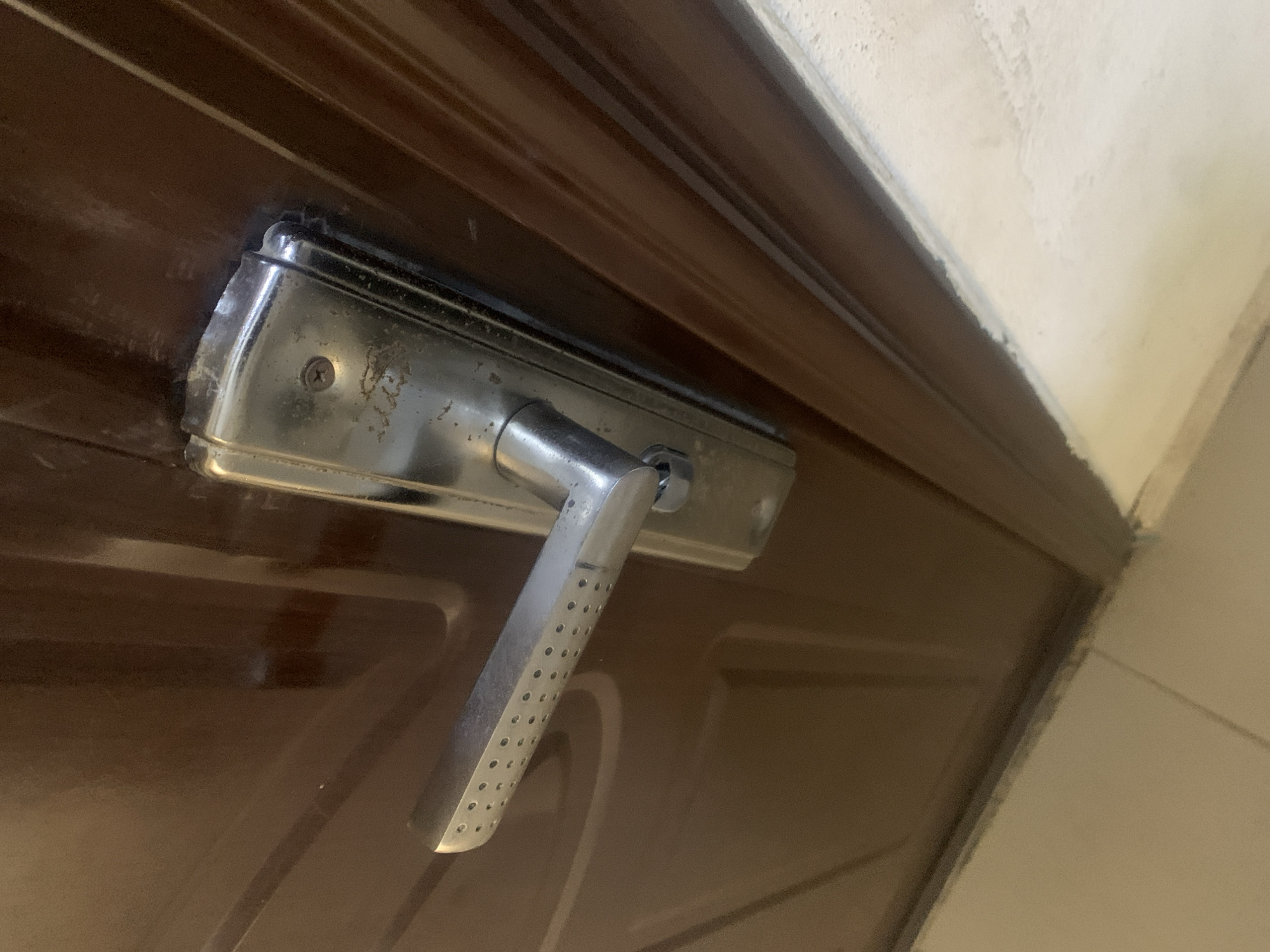 The image shows a close-up view of a metallic door handle and lock mechanism on a brown, polished wooden door. The handle is silver-toned with a textured grip, featuring a series of small circular perforations. It is attached to a rectangular plate that houses the lock, which has a visible keyhole. The door is part of a larger frame, with a section of a lighter-colored wall visible to the right, suggesting an interior setting. The lighting appears to be artificial, casting reflections on the polished surfaces of the door and handle, indicating it might be indoors. The image does not depict any people or specific activities. The overall impression is one of a functional, everyday object, likely a residential or commercial door, given the simple design of the hardware. There is no visible text in the image. The provided context of Jalingo, Nigeria, places the scene geographically, but offers no specific visual cues within the image itself to confirm this location.