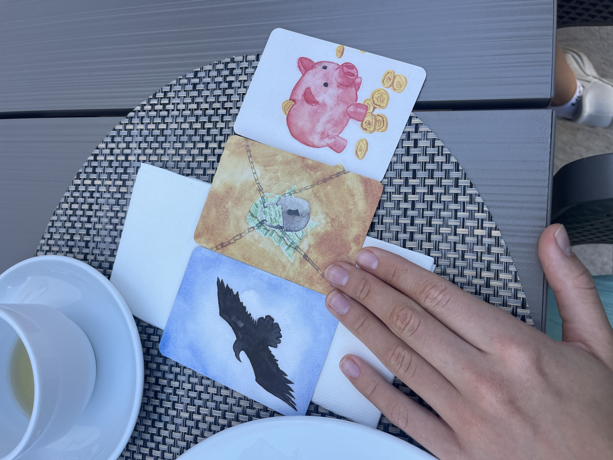 The image depicts an outdoor table scene in Budva, Montenegro. A person's bare left hand is visible in the lower-central portion of the frame, resting on a stack of white napkins. To the upper left, a white ceramic teacup containing a light yellow liquid sits on a matching saucer. Three distinct, hand-painted cards are horizontally arranged on a dark and light grey woven placemat.
The leftmost card displays a black raven in flight against a light blue background. The central card shows a stack of green bills secured by a padlock and chain, set against a light brown backdrop. The rightmost card features a pink piggy bank surrounded by gold coins, with one coin entering the bank's slot, on a white background. The placemat rests on a dark grey outdoor table with a distinct ribbed texture, visible in the upper and lower right segments. A vertical seam separates two sections of the table surface. A dark, possibly metal, chair leg is partially visible in the lower-left corner. Below the table in the lower right, a person's left foot, clad in a white sneaker with a dark sole, is discernible.