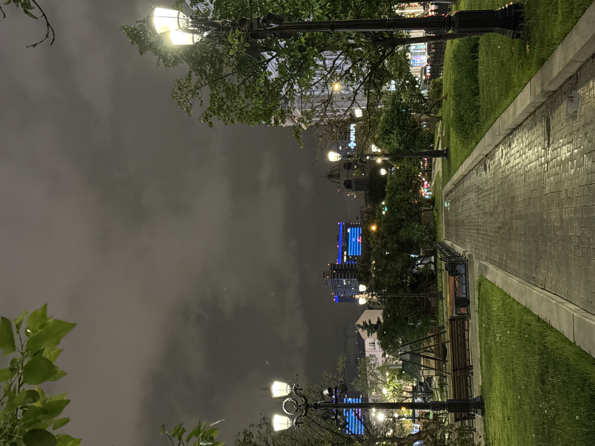 The image depicts a nighttime scene in Moscow, Russia, featuring a pathway illuminated by streetlights lining a park. A brick path stretches into the distance. To the sides, there is grass and greenery. Buildings are visible in the background, some with illuminated signs and blue lighting. The sky is cloudy. Benches are along the pathway.